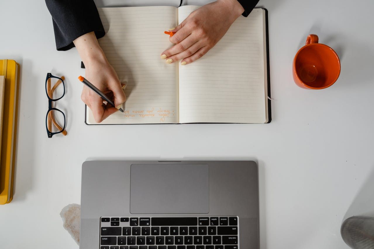  A person writing notes in an open notebook beside a laptop, an orange mug, and glasses.