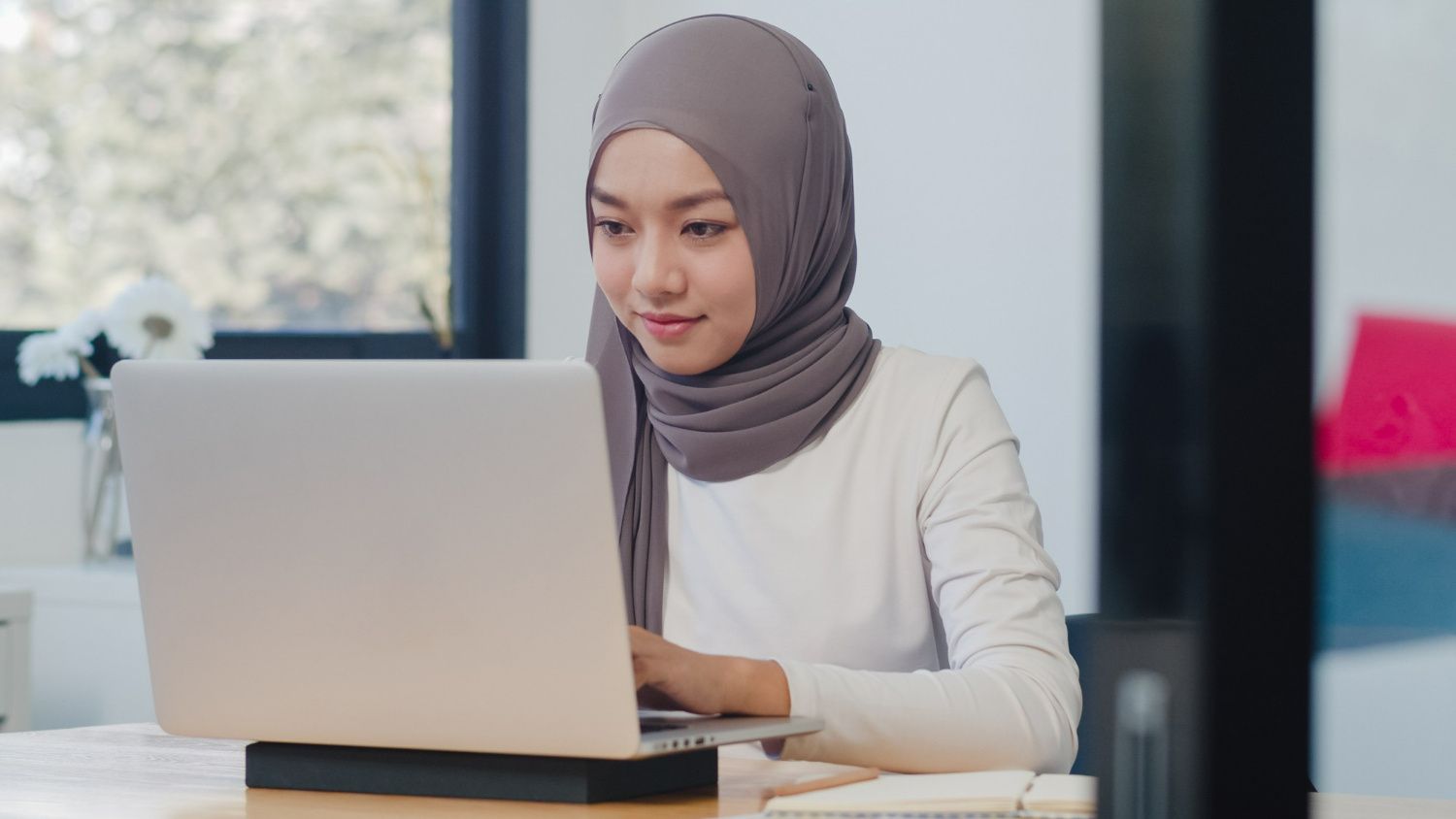 Young Muslim woman in a hijab working confidently on a laptop at a modern office desk with natural light, while researching a job verification letter example. 