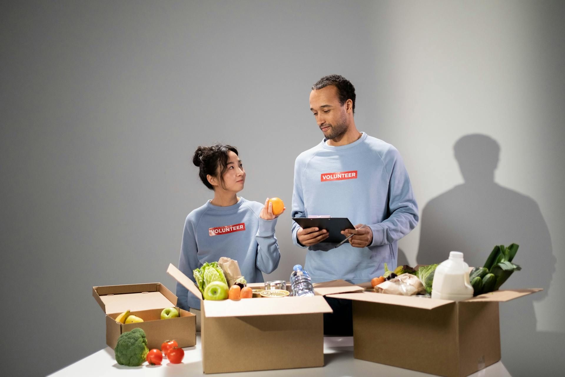 Two volunteers in matching light-blue shirts pack food boxes, smiling and talking while sorting fruits and vegetables.