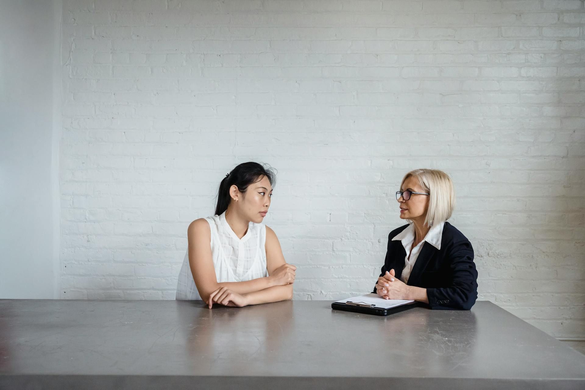 A woman and another woman sit across from each other at a table, having a conversation, with a folder placed in front of one of them.