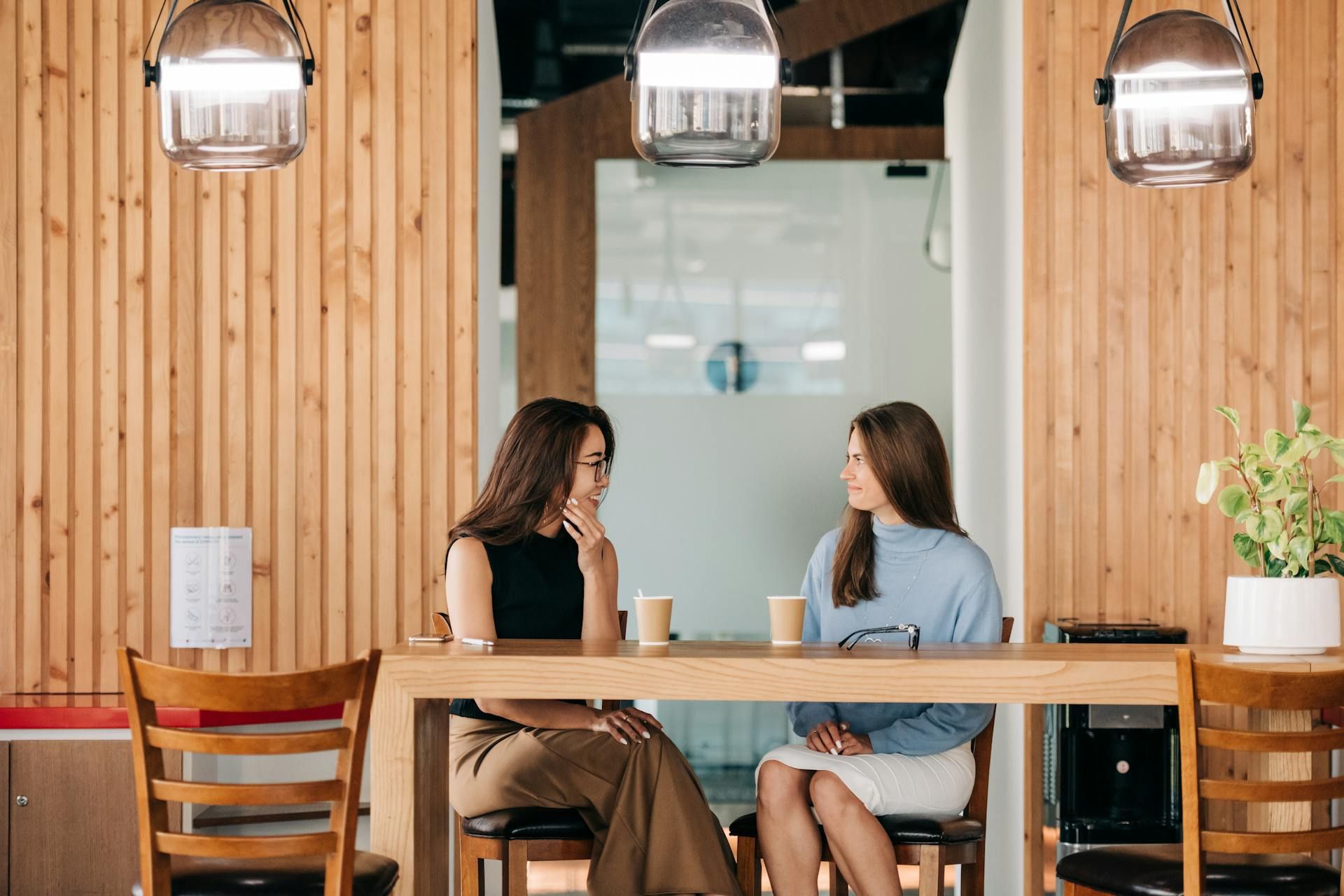 Two women chatting over coffee at a wooden table in a modern café with hanging lights and warm wooden walls.