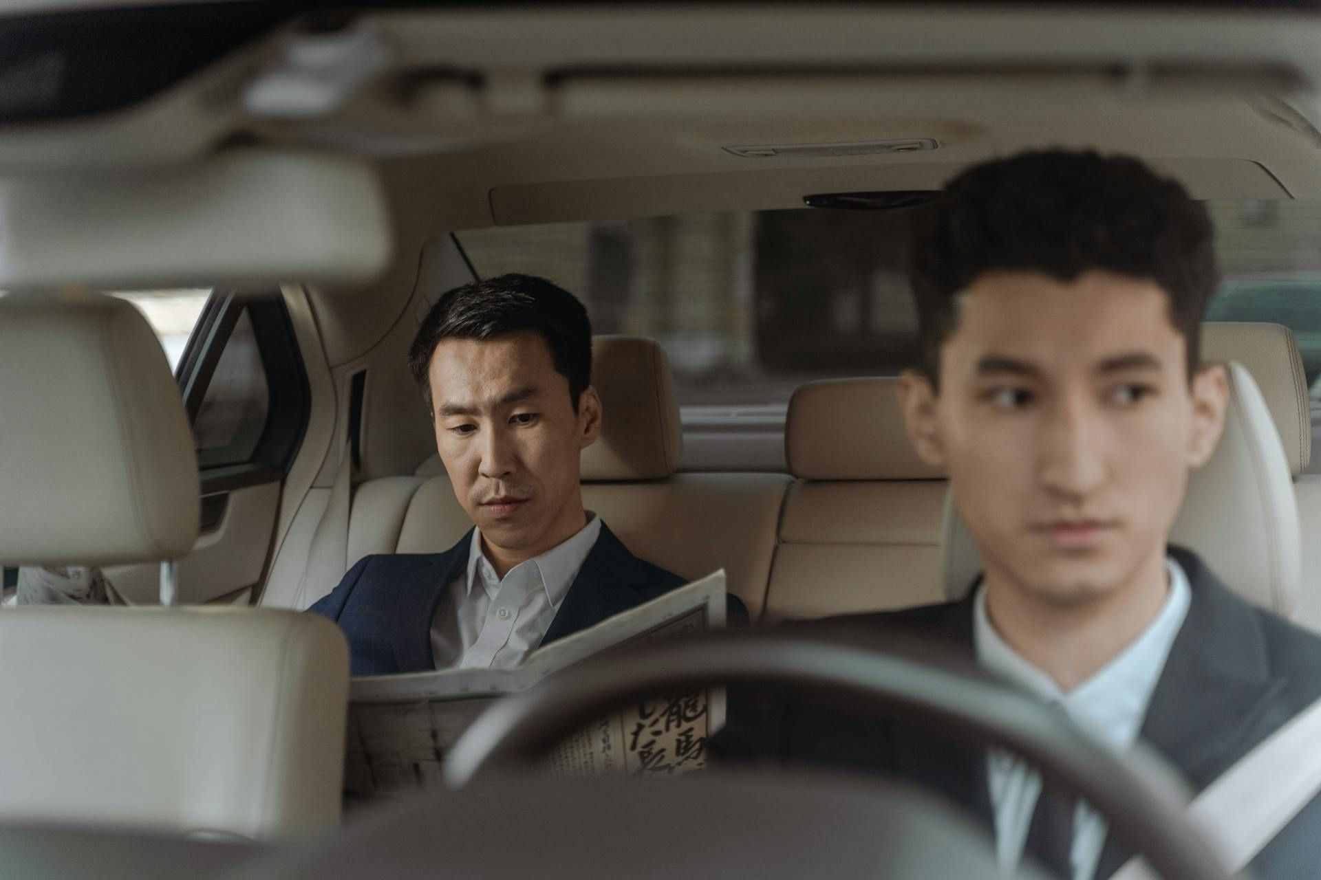 A man reads a newspaper in the back seat while another man drives, both in formal attire inside a clean, modern car.