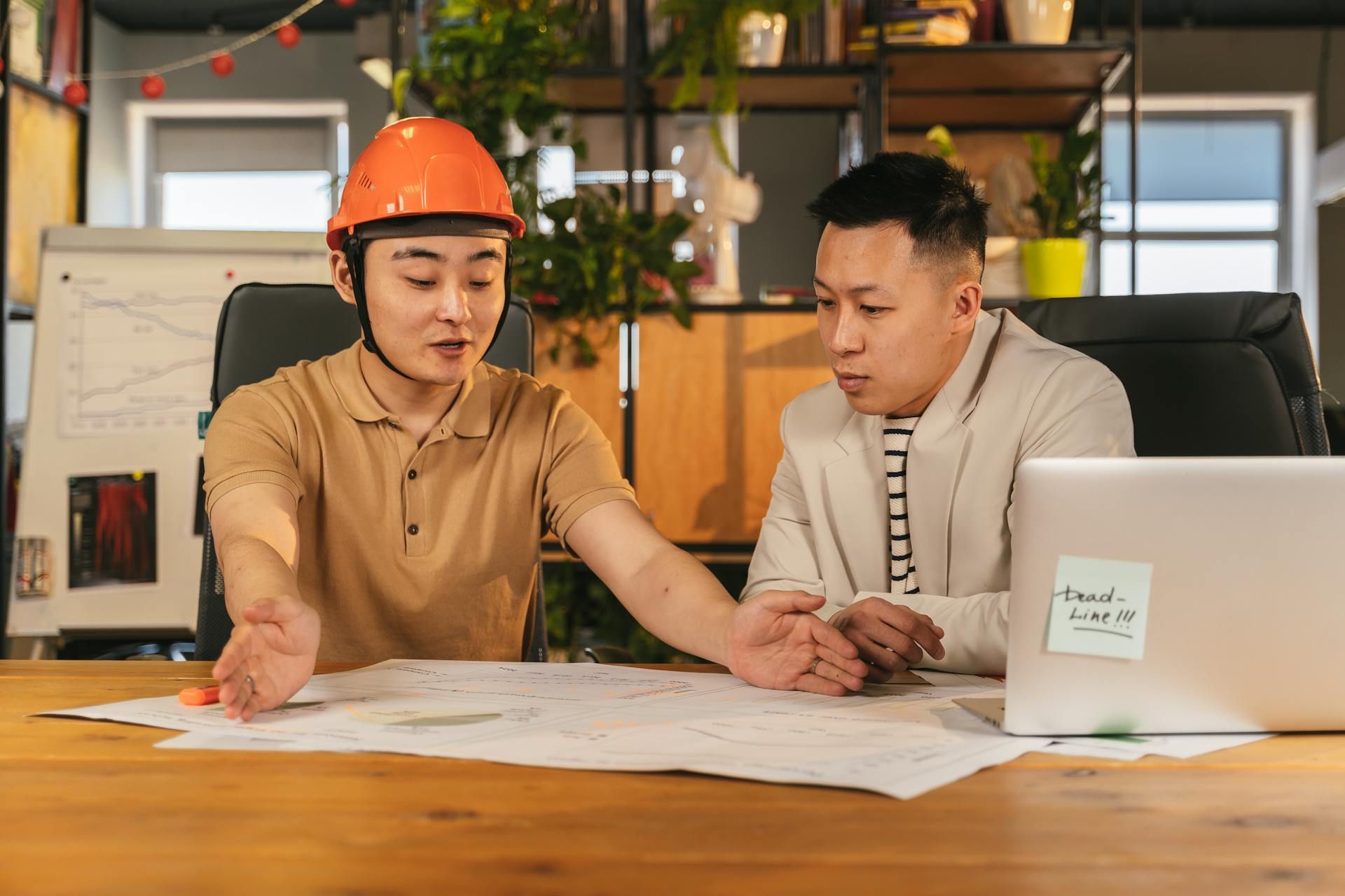 A man in a safety helmet explains plans on a table while another man in a blazer reviews the drawings and listens attentively.