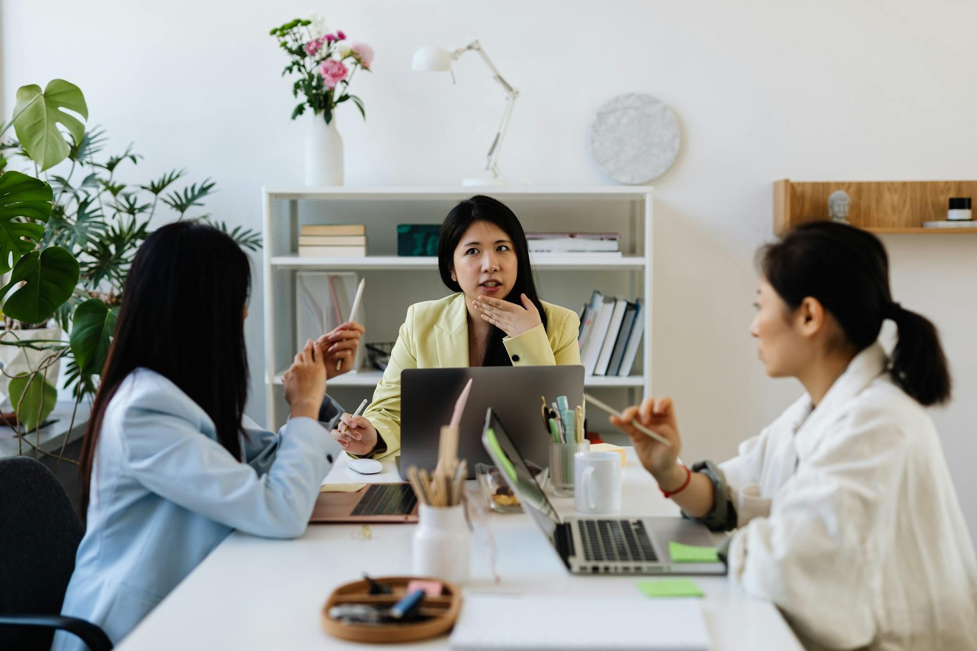 Three women discussing around a table, using laptops and notes in a bright modern office. 