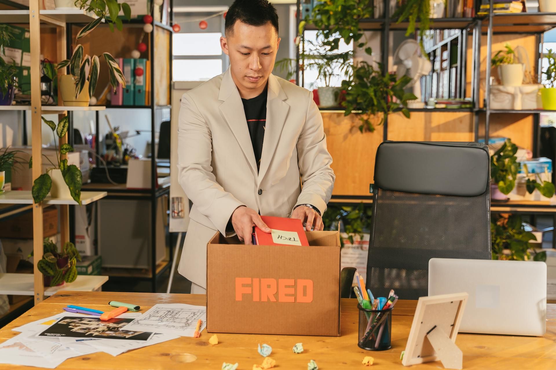 Man packing personal belongings into a cardboard box labelled “Fired” in a modern office setting.