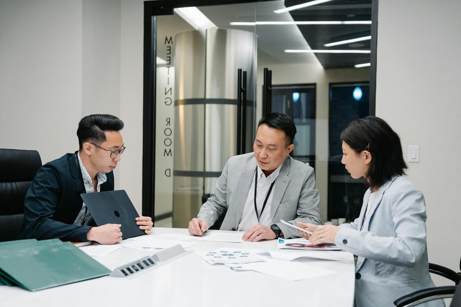 Three professionals reviewing business documents during a strategy meeting in a corporate boardroom.