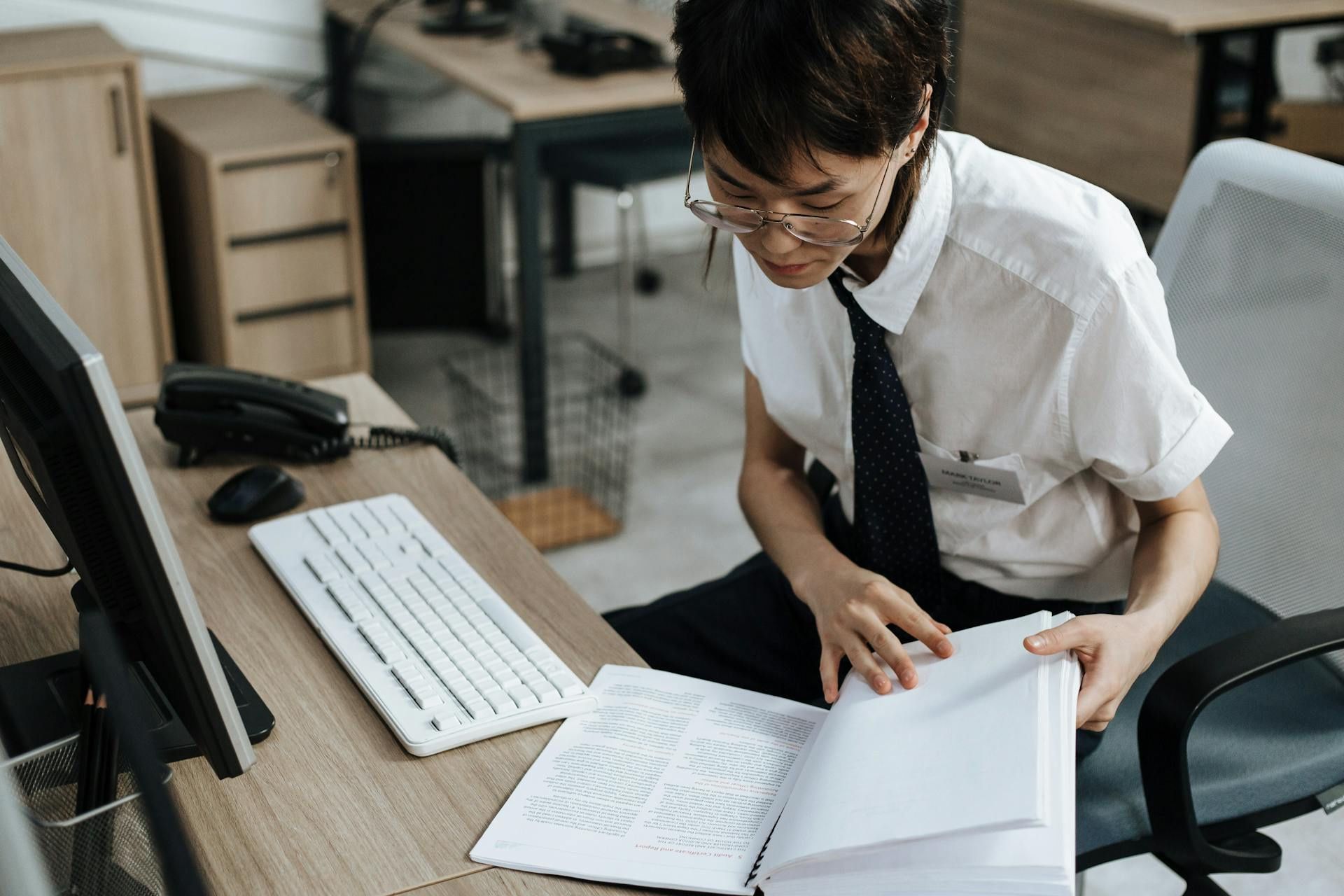 Focused employee reading a printed manual at a desk with a computer and phone.