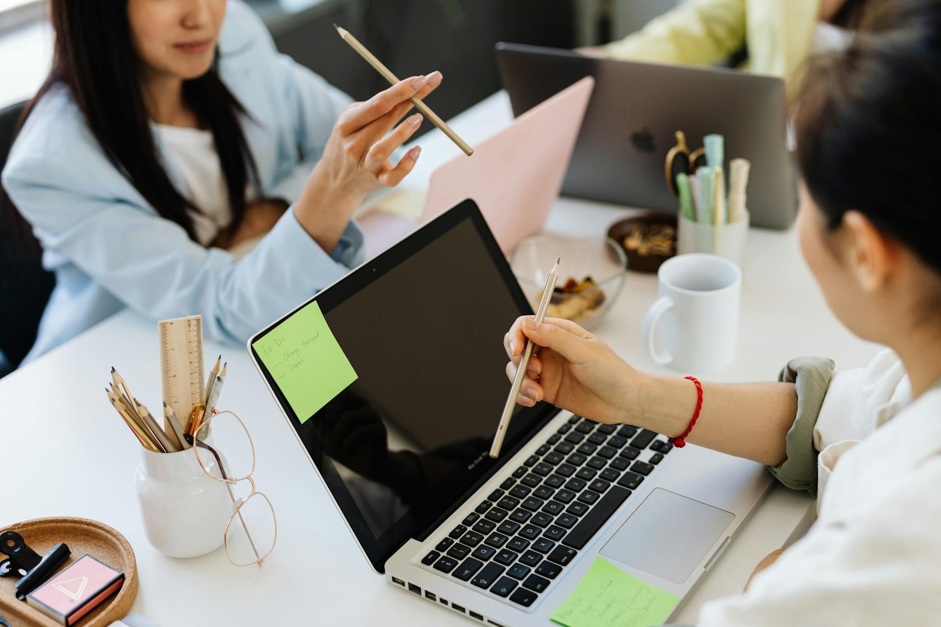 Close-up of colleagues using pencils to discuss ideas around a laptop with sticky notes and office supplies.