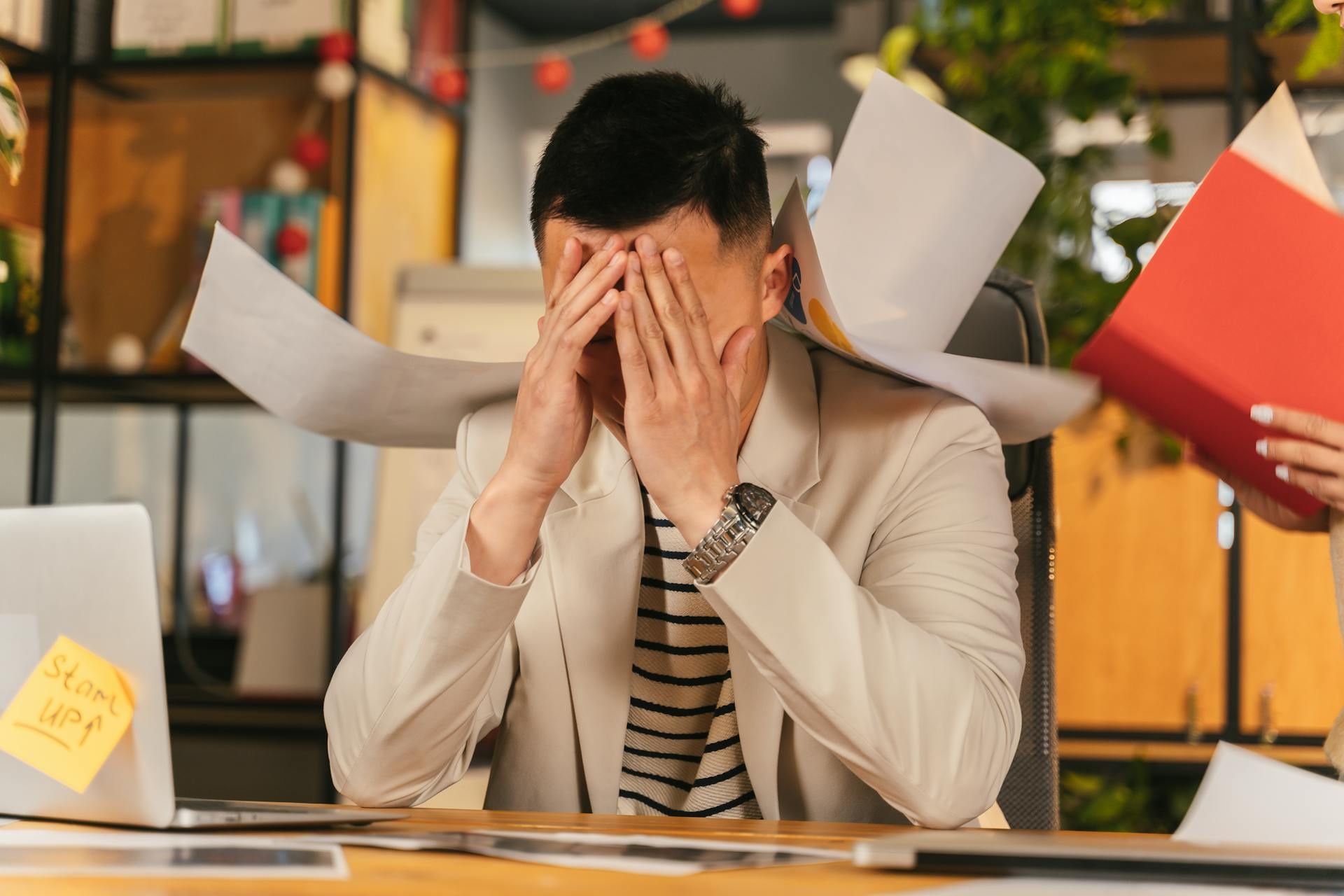 A stressed man sits at his desk with his hands covering his face as papers fly around him.