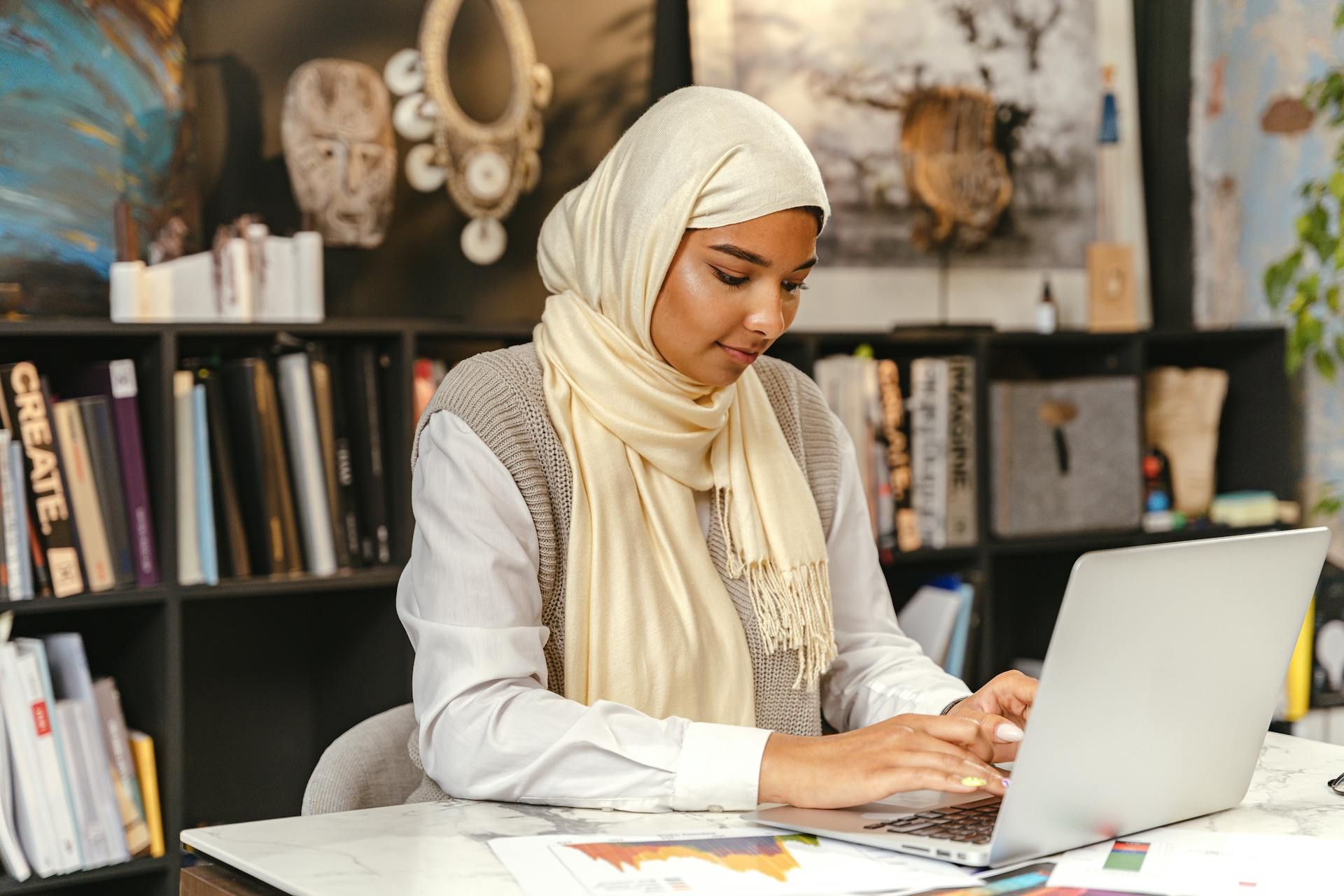 Seorang wanita bertudung bekerja menggunakan komputer riba di meja, dengan buku dan hiasan seni di latar belakang.