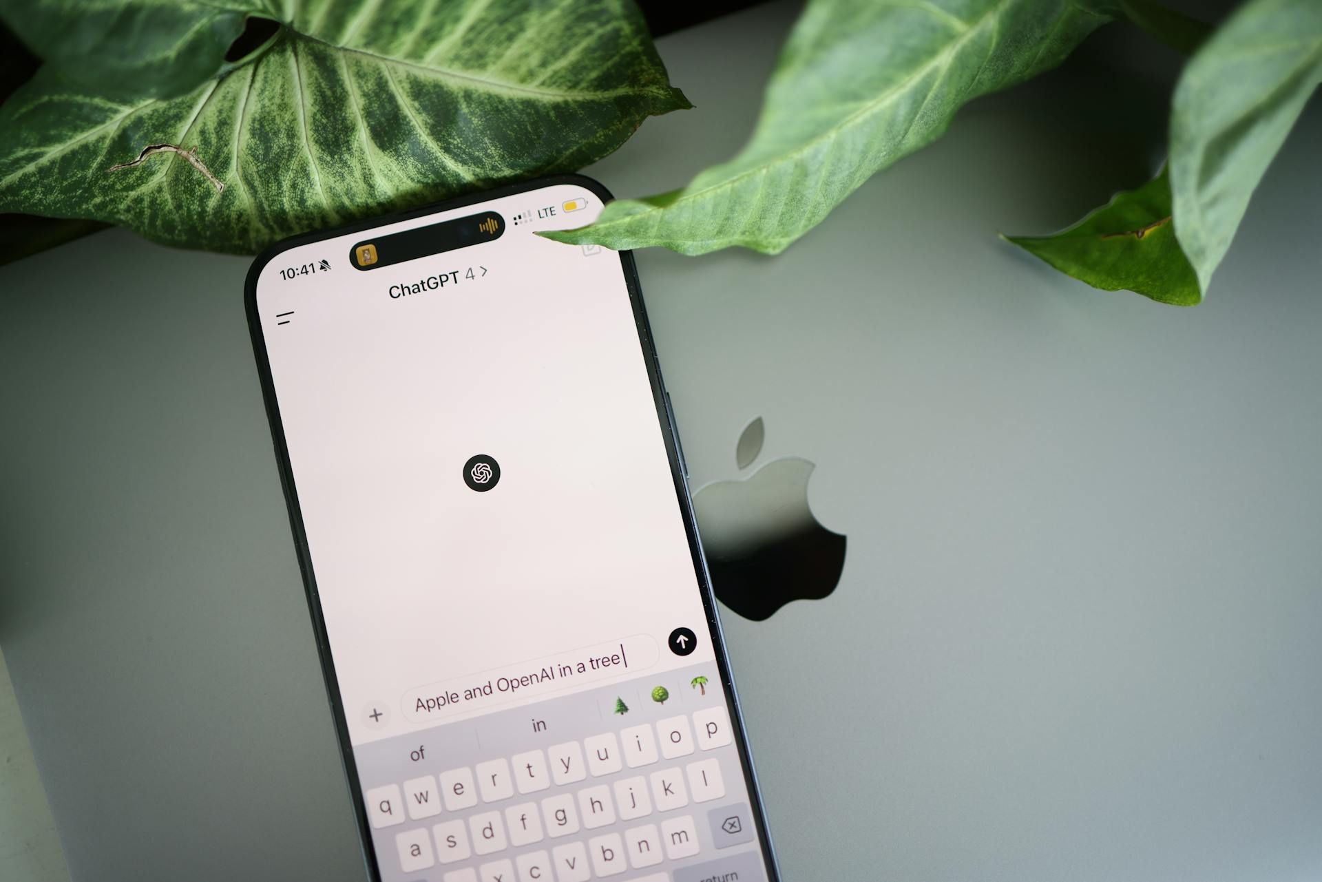 A smartphone displaying the ChatGPT app rests on a MacBook beside green leaves.