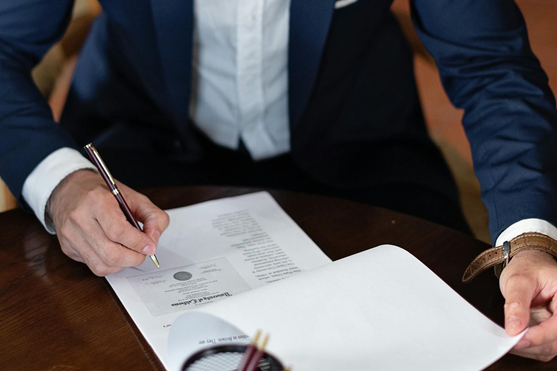 A person in a navy suit is signing a document on a wooden table, holding a pen and reviewing paperwork in a formal setting.