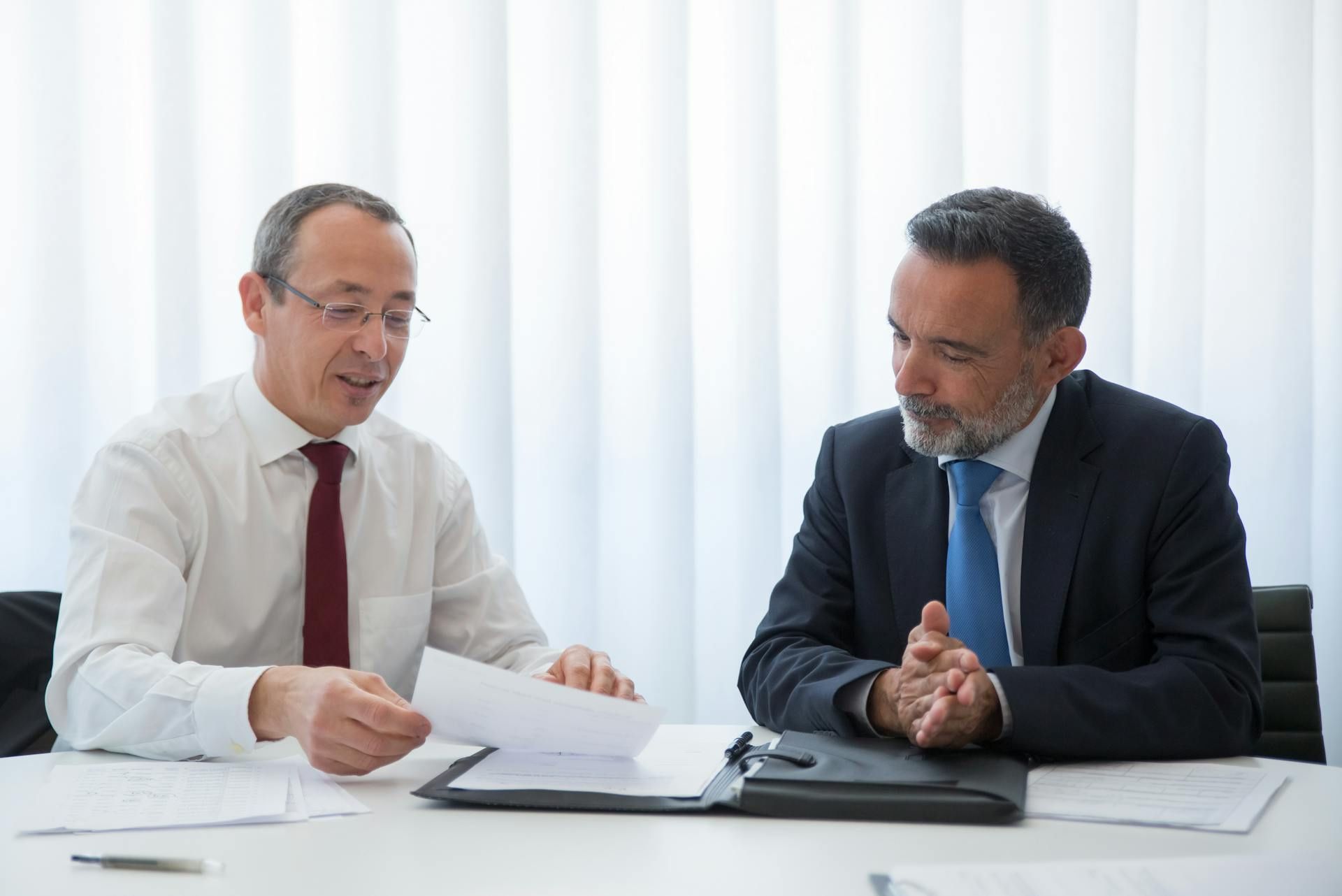 Two businessmen in formal attire discussing documents at a desk, appearing engaged in a professional meeting.