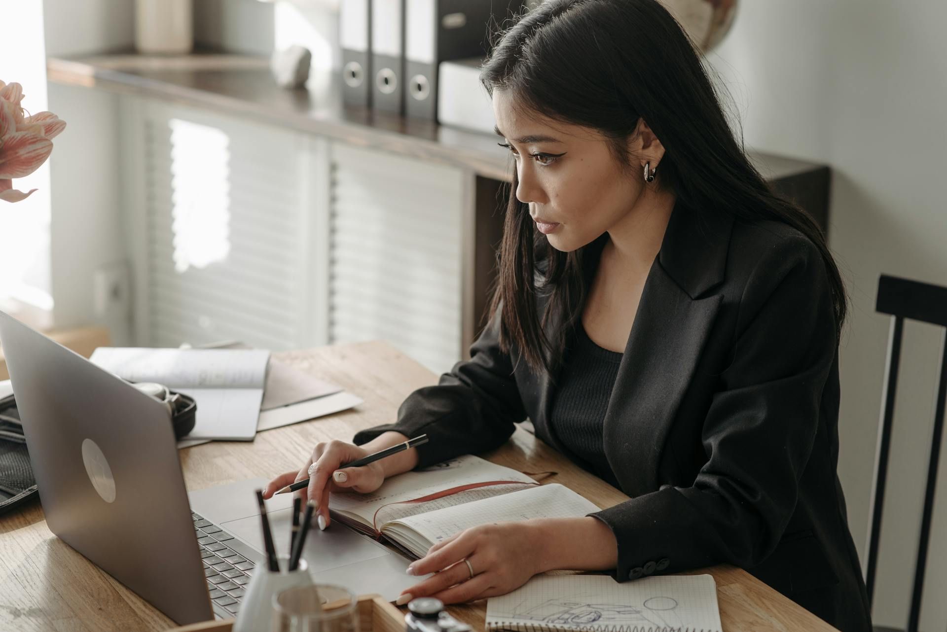A woman working at a laptop while writing in a notebook in a home office setting.