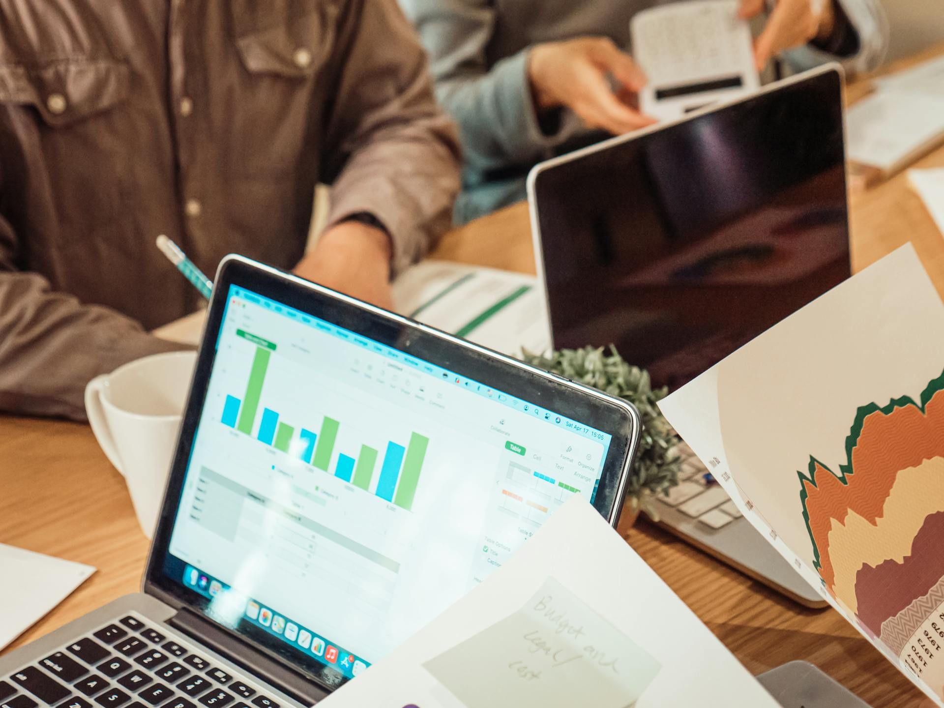Beberapa individu sedang berbincang di meja dengan komputer riba dan carta kewangan yang dipaparkan, meneliti graf bar dan dokumen analisis data.
