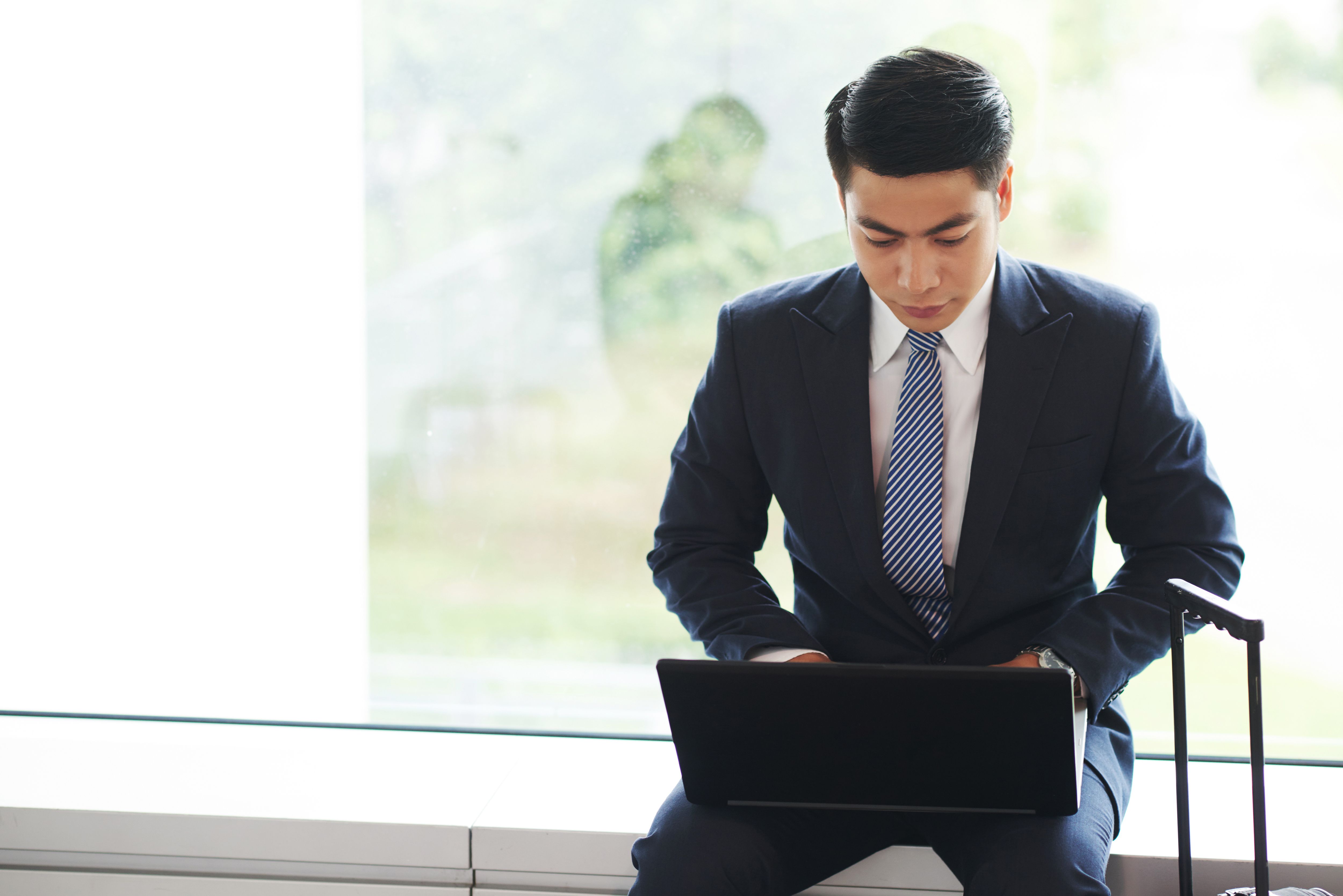 Businessman in a suit sitting by a window with a suitcase beside him, working on a laptop—possibly reviewing or composing a professional email format.