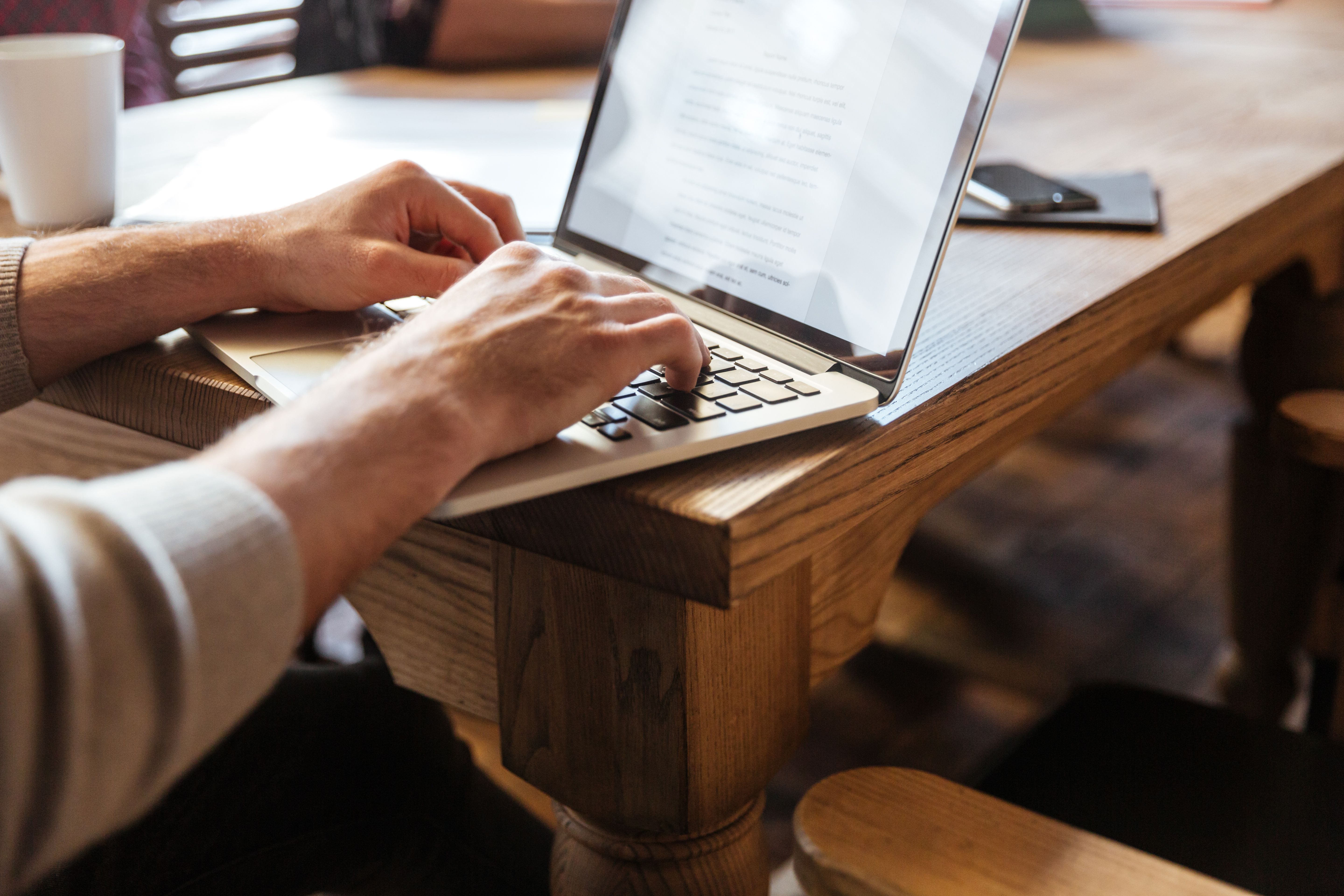 Close-up of a person typing a formal letter format on a laptop,  with documents, coffee, and phone on a wooden desk. 