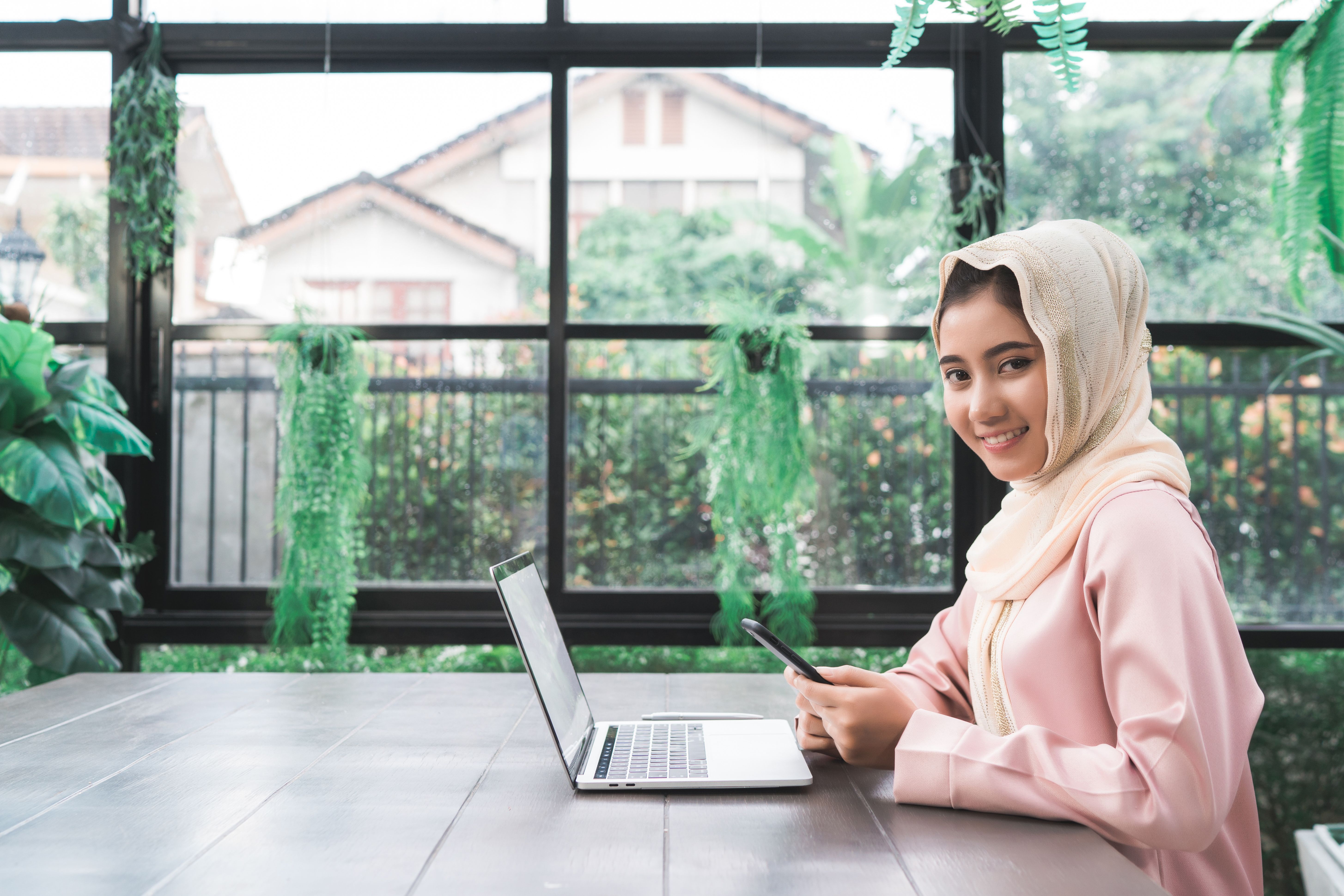 Smiling young woman working on a laptop and holding a smartphone in a bright indoor garden setting, possibly reviewing email format for professional communication.