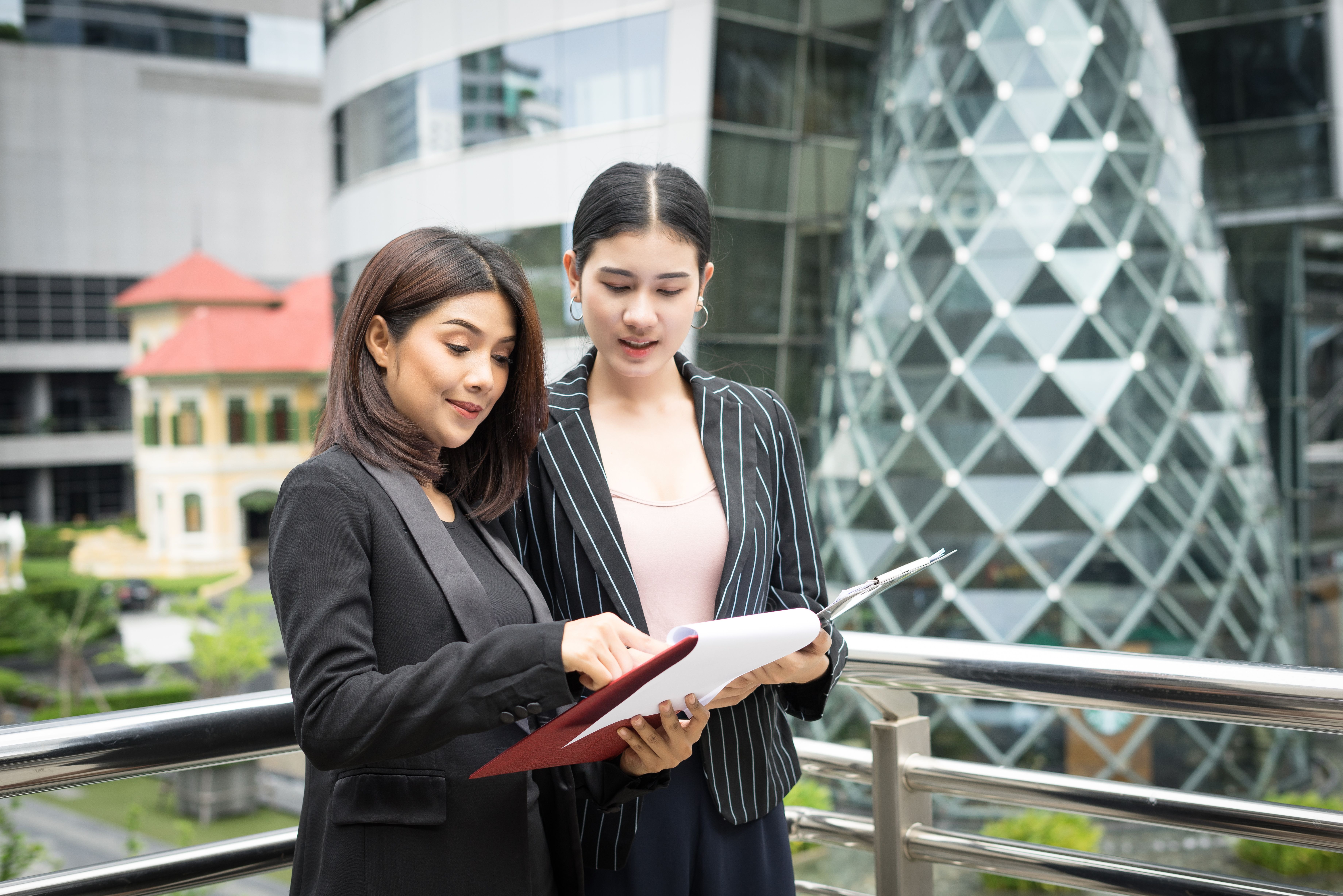 Dua wanita berpakaian formal sedang berdiri di luar bangunan pejabat moden sambil meneliti dokumen di dalam fail, menunjukkan perbincangan kerja atau semakan tugasan.