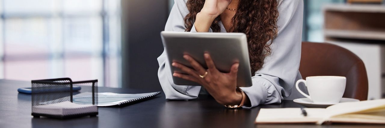 woman at a home office with notepad and coffee cup reviewing a tablet