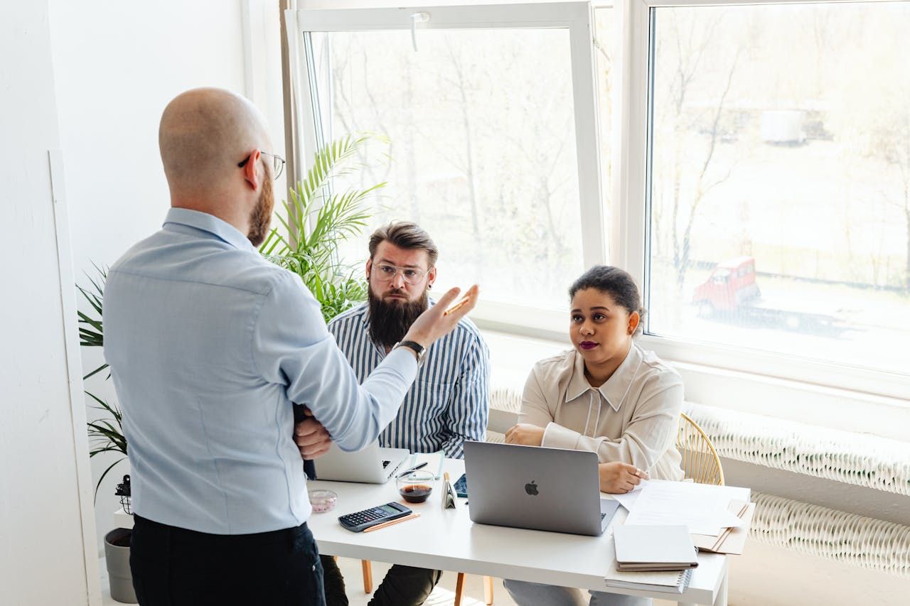 Seorang mentor berkepala botak memberi penerangan kepada dua mentee yang mendengar dengan fokus di meja kerja mereka dalam suasana pejabat moden yang terang.
