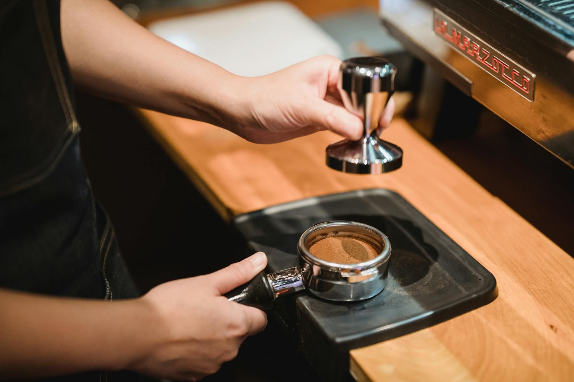 Barista sedang memadatkan serbuk kopi ke dalam penapis menggunakan alat penekan kopi di sebuah kedai kopi.