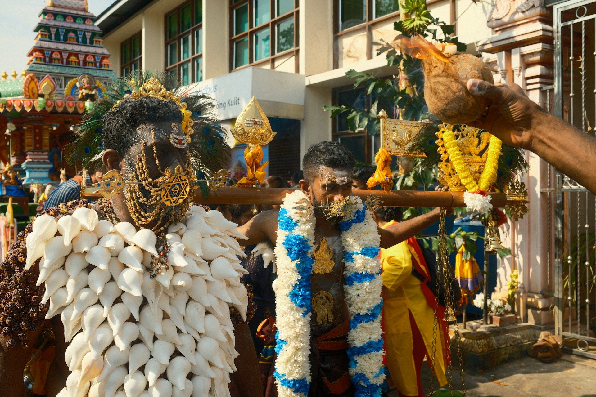 Dua lelaki mengambil bahagian dalam perarakan Thaipusam, mengenakan pakaian tradisional, kavadi berhias dan tusukan muka, diiringi asap dan suasana keagamaan di hadapan kuil.