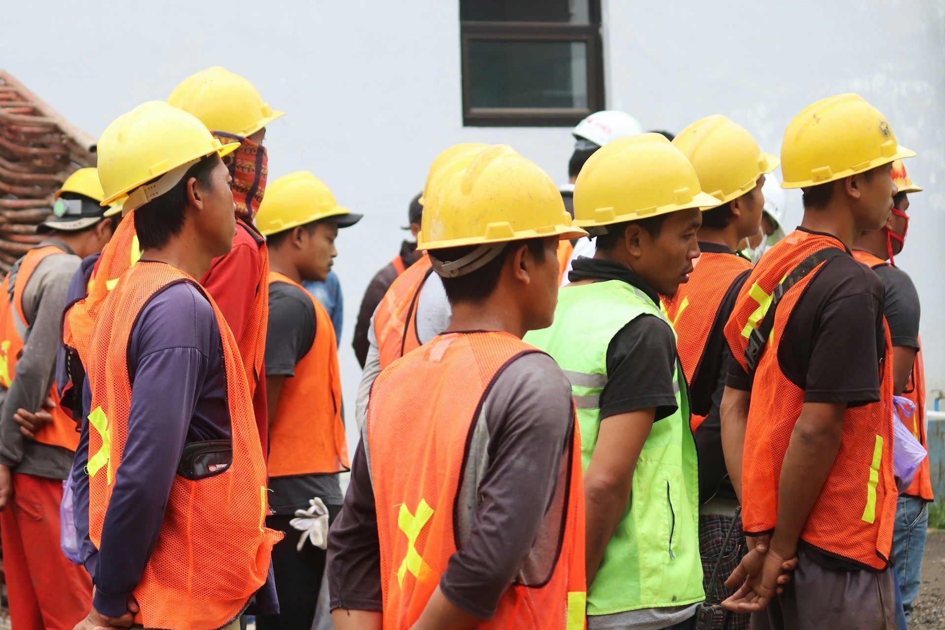 Sekumpulan pekerja asing memakai topi keselamatan dan vest keselamatan berkumpul di tapak pembinaan untuk taklimat kerja.