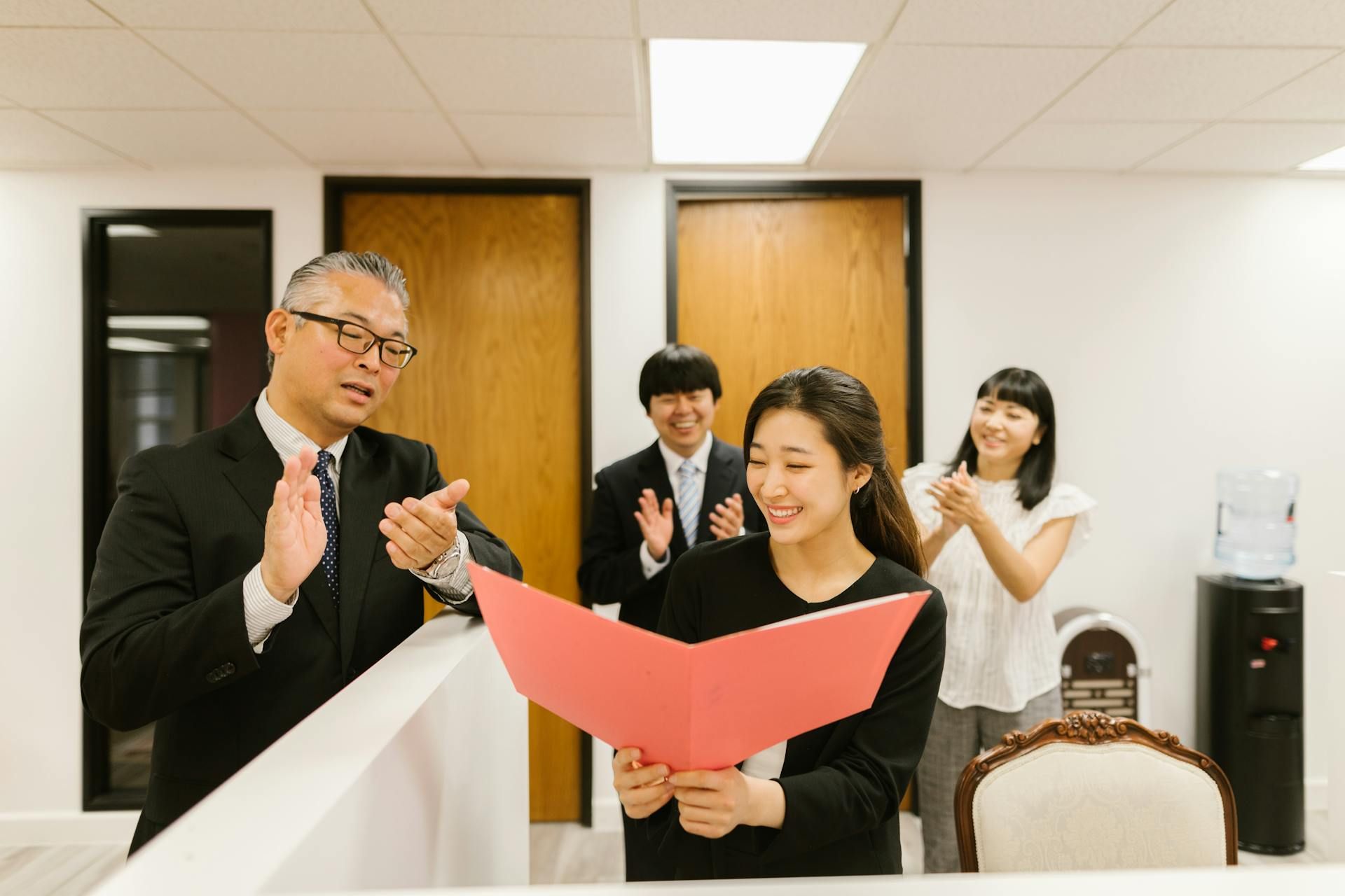 A team of colleagues clapping and congratulating a woman holding a red folder in an office setting.