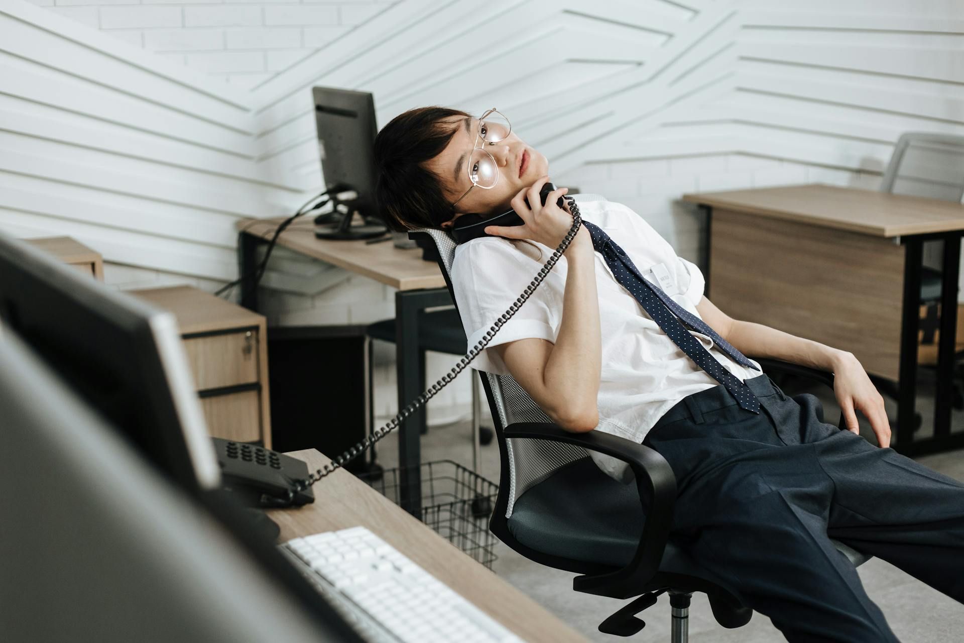 A man sitting in an office chair with a relaxed posture, holding a phone to his ear in a minimalist office setting.