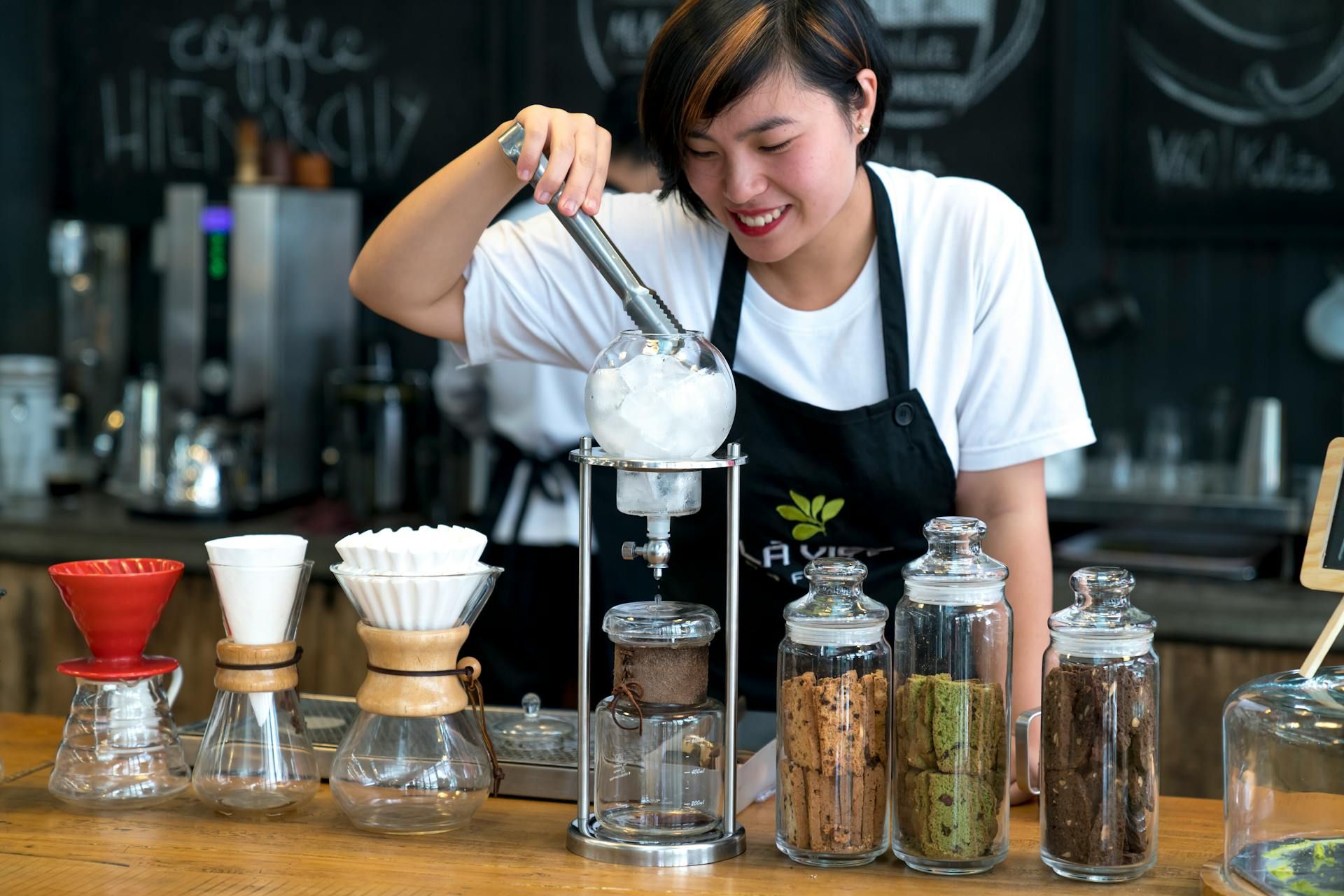 Barista wanita menyediakan minuman di kafe, dikelilingi oleh bekas berisi biskut dan peralatan kopi.