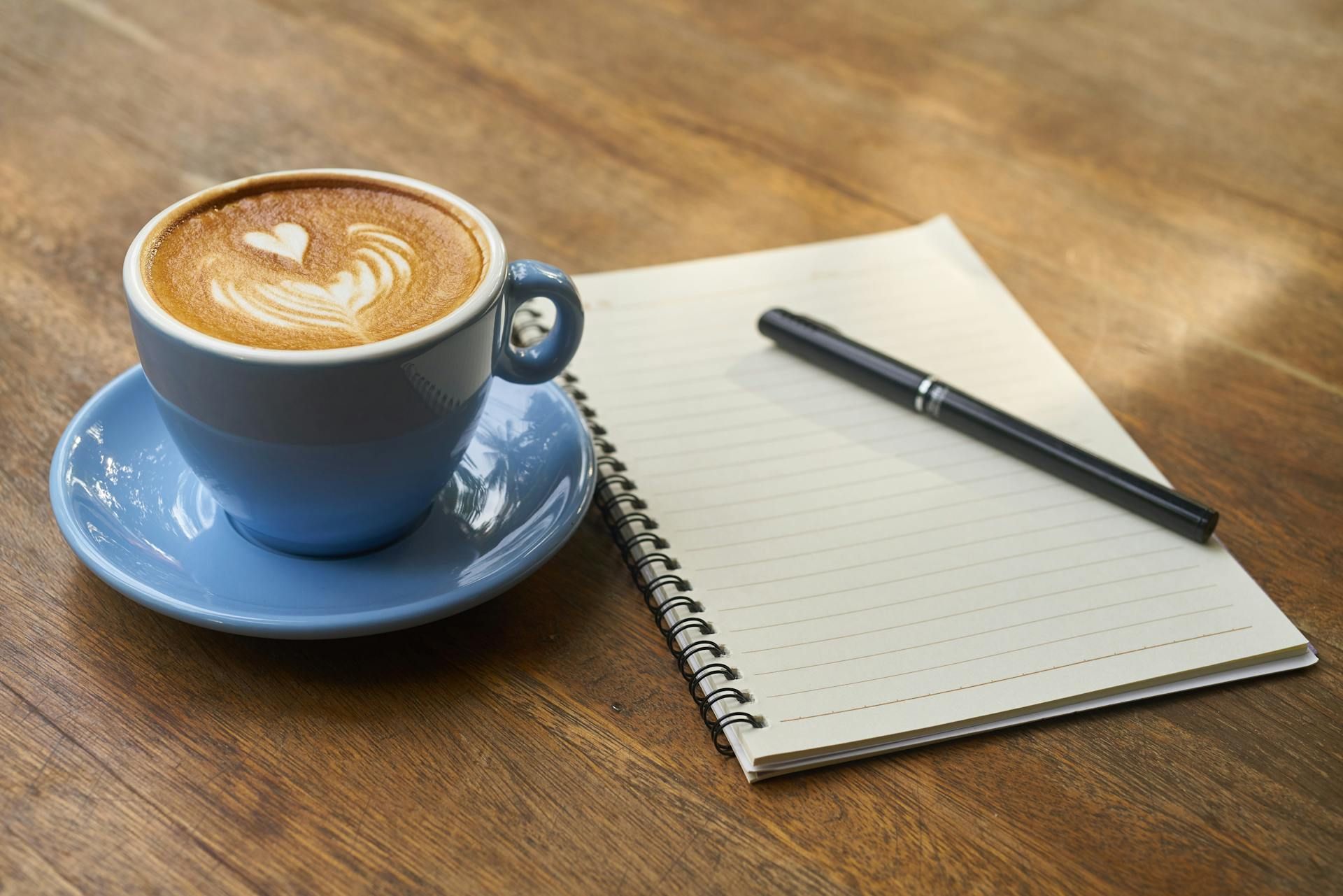 Secawan kopi latte di atas meja kayu dengan buku nota kosong dan pen hitam di sebelah.