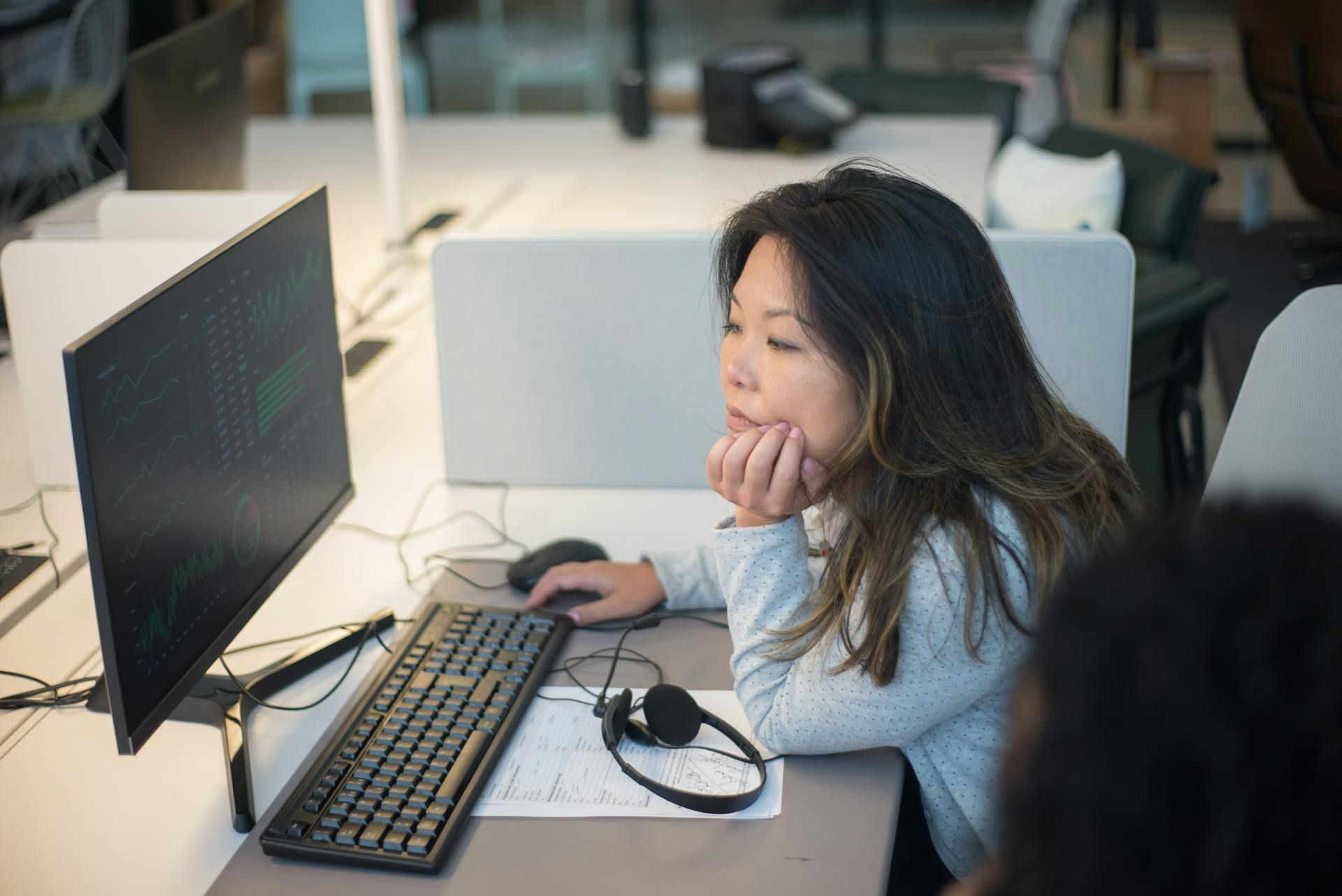 A woman sitting at a desk, resting her chin on her hand as she stares at the computer screen, wearing a headset in a modern office environment.