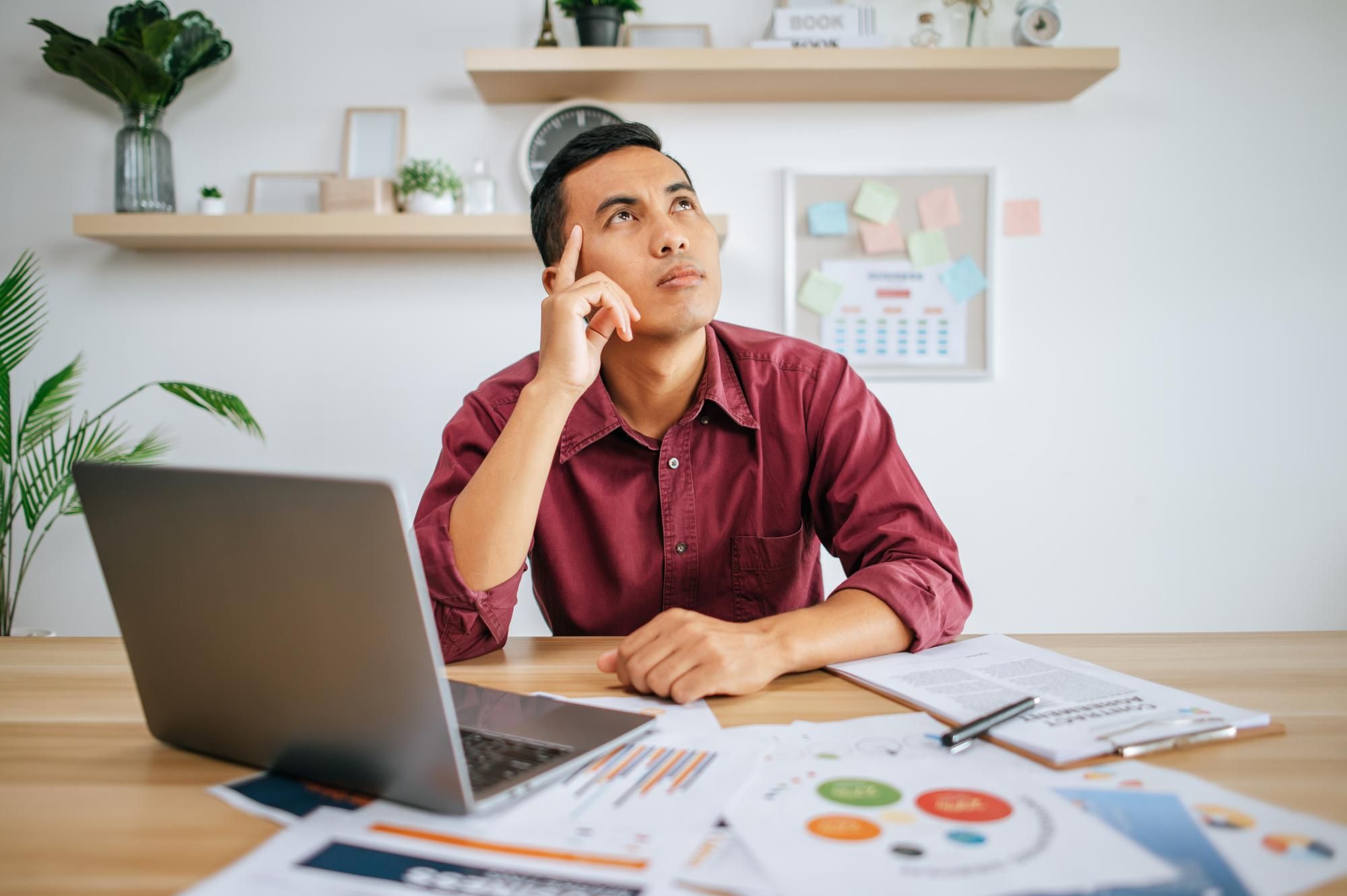 Man in a red shirt sitting at a desk with charts and a laptop, thinking about employee performance evaluation examples. 