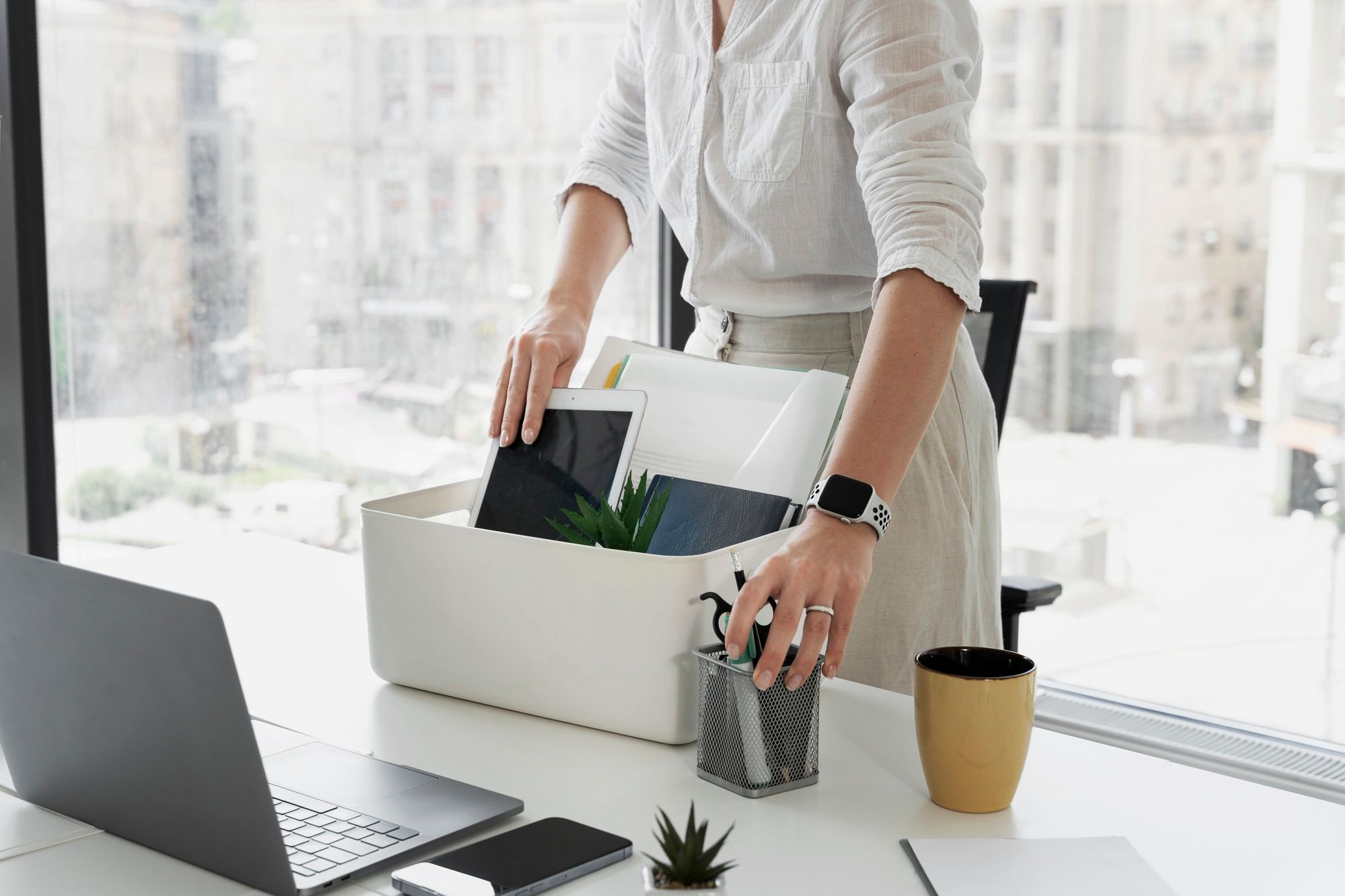 Person standing at a desk and packing office items into a box. This suggests they are resigning or leaving their work and ready to start their job search.