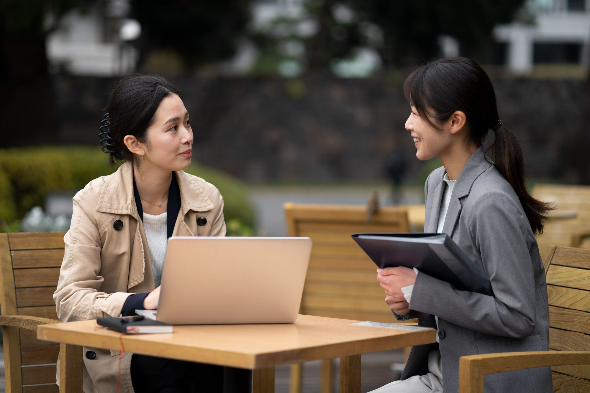Two women in business attire have a professional conversation outdoors, one with a laptop and the other holding a folder.