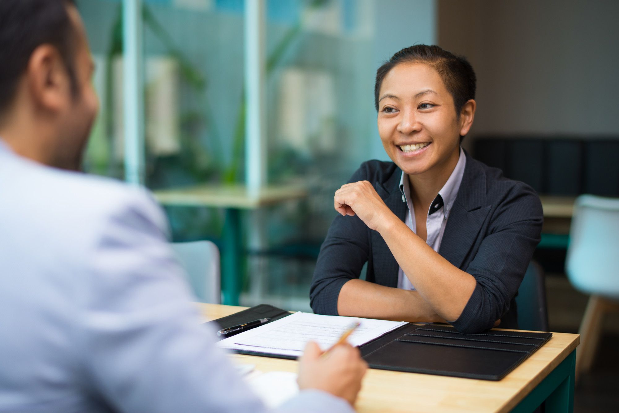 A smiling businesswoman in a blazer sits at a desk during a job interview, engaging confidently with the hiring manager.