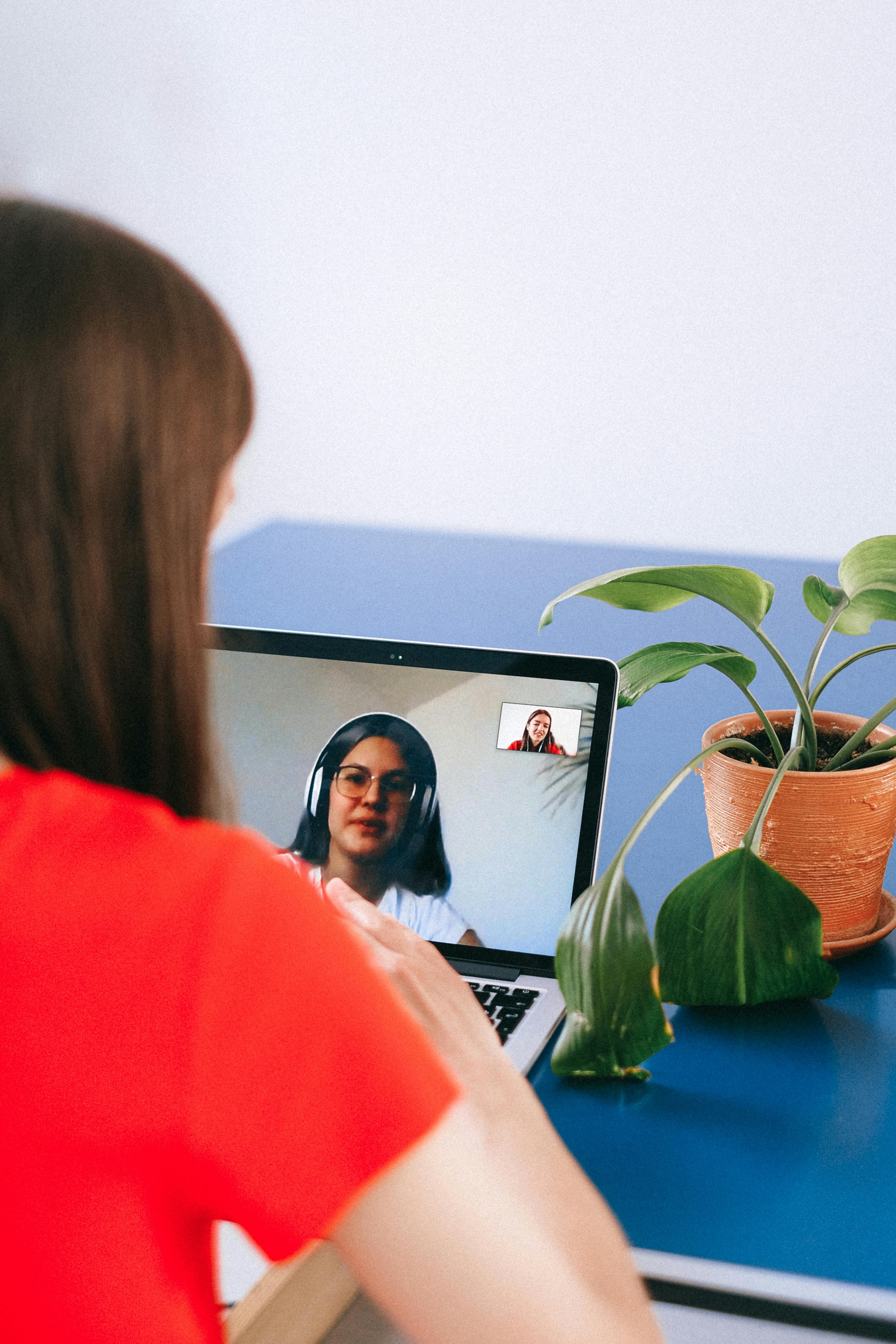 A person in a red shirt in a video call, speaking with another woman wearing headphones and glasses.