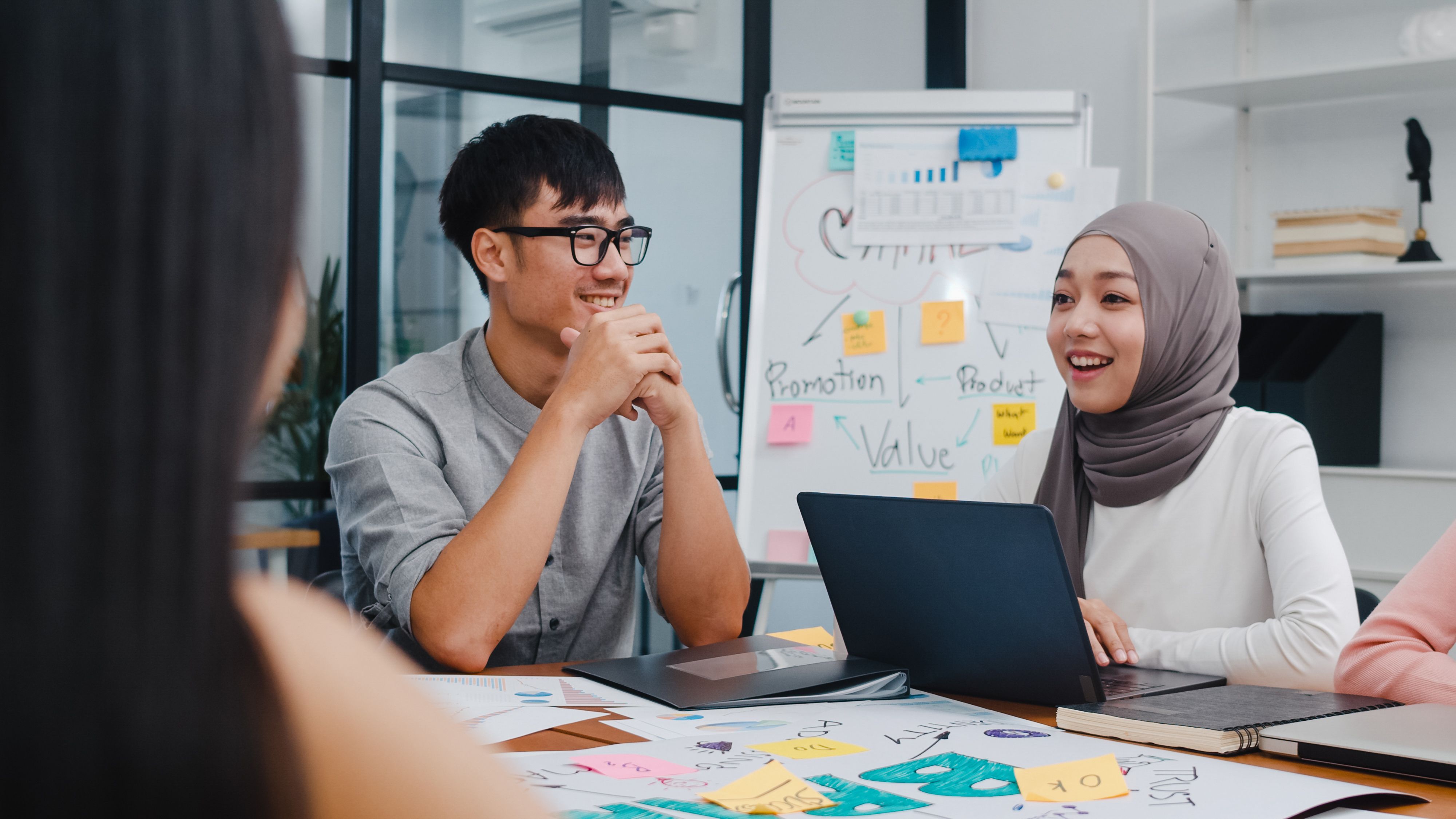 Seorang perempuan yang memakai tudung bersama dengan seorang lelaki memakai cermin mata sedang berbincang di dalam pejabat.