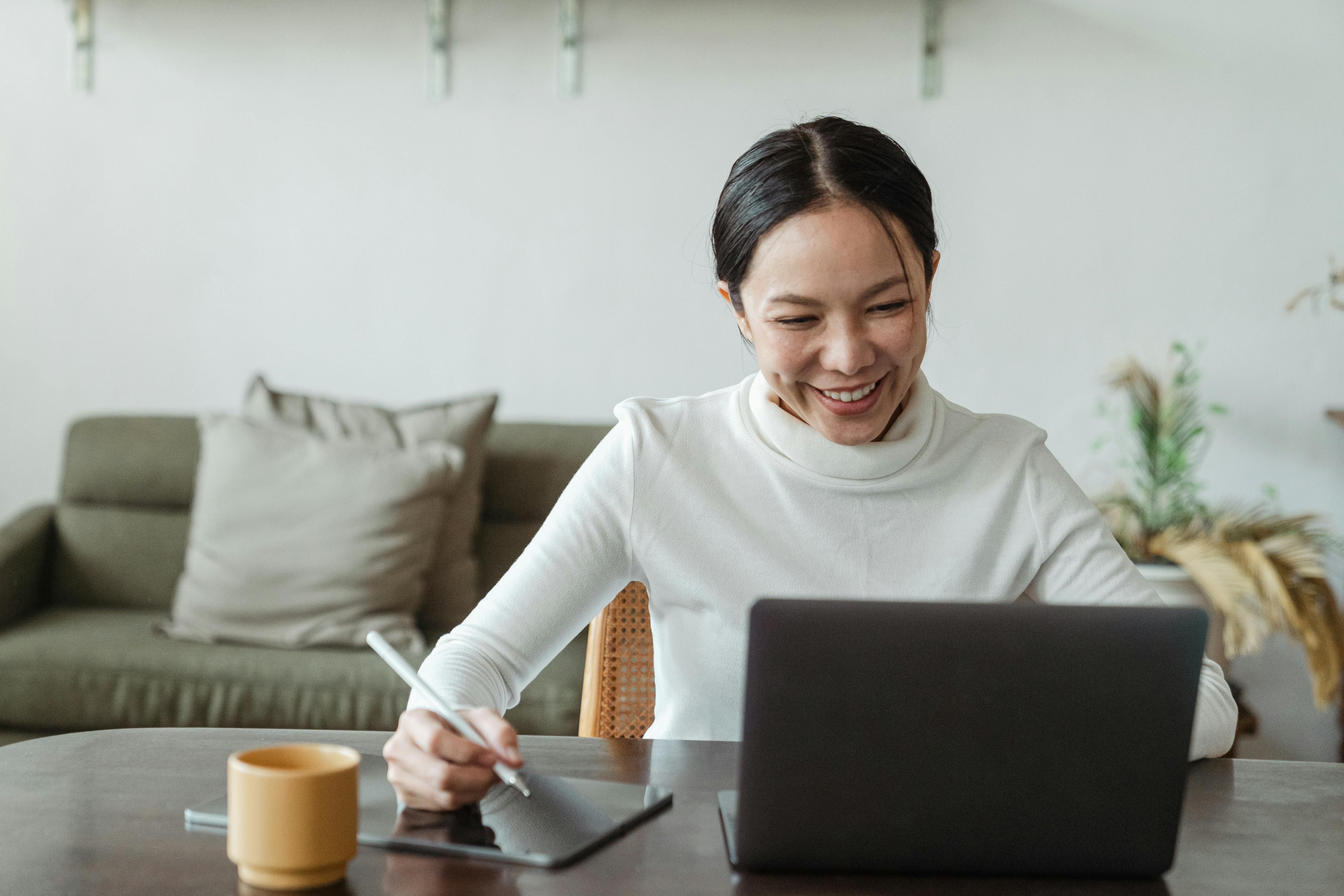 A smiling woman using a digital pen tablet while working on a laptop at home, seated at a table with a yellow coffee cup.