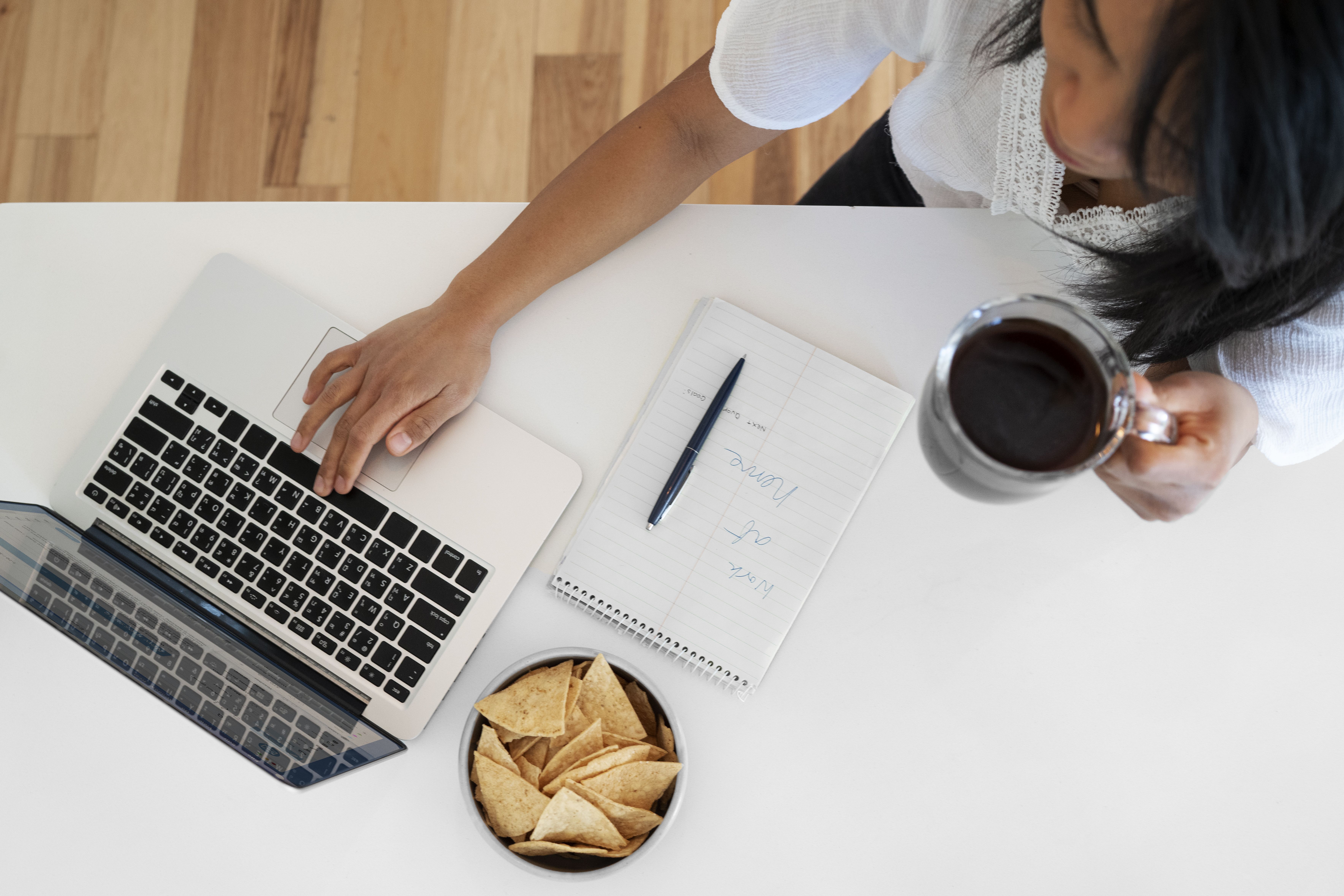 Top view of a woman using a laptop and holding a cup of coffee while drafting a follow up email after interview, with notes and snacks on the table. 