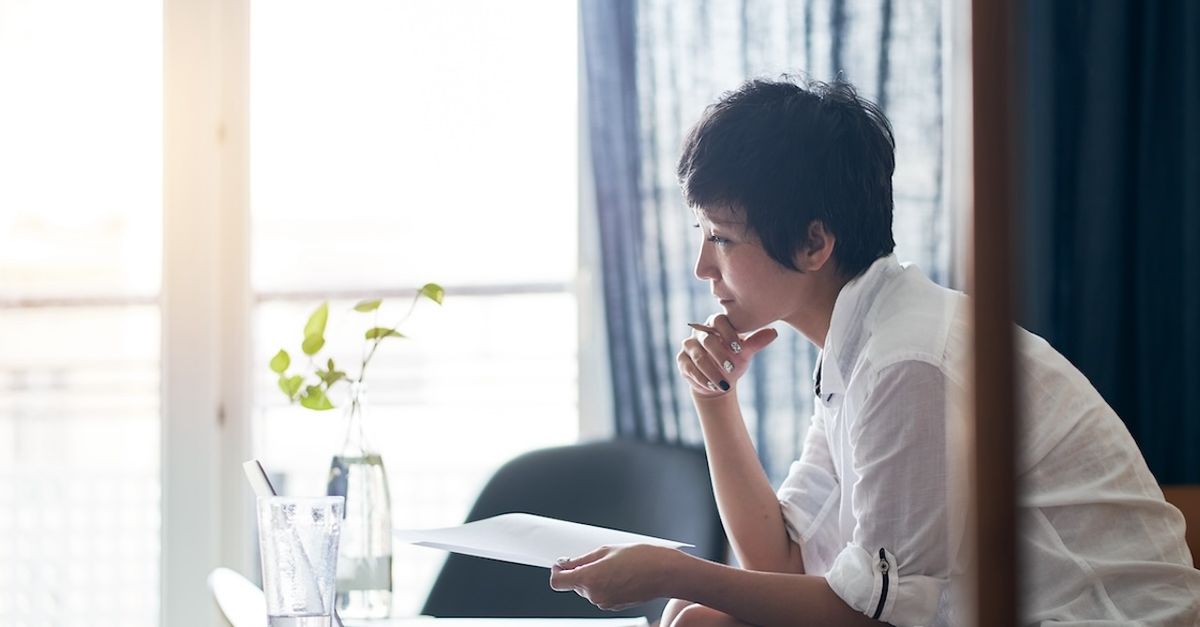 a woman writing with a pen and piece of paper, looking off in deep thought while holding her chin