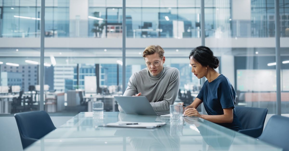 a woman and man sit at a work table looking at a laptop, with two glasses of water and a dry-erase nearby