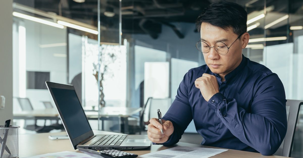 a man in a dark blue shirt working at a desk in an office, with a laptop next to him